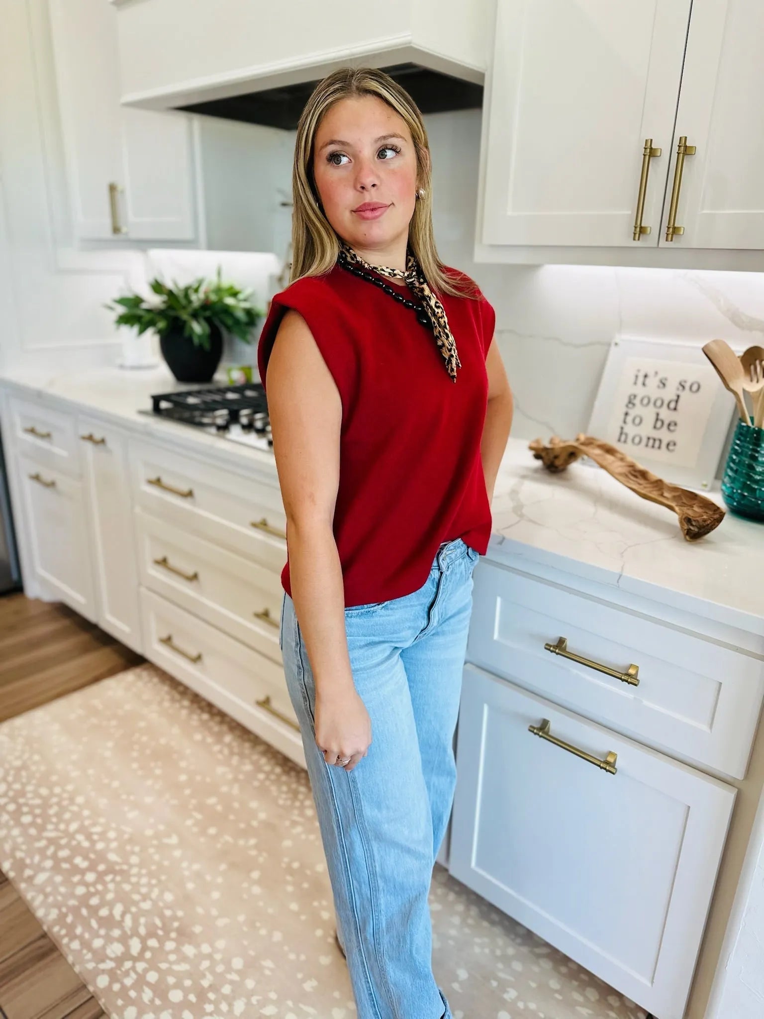 Woman in red round neck cap sleeve sweater and light wash jeans standing in a kitchen, accessorized with a leopard print