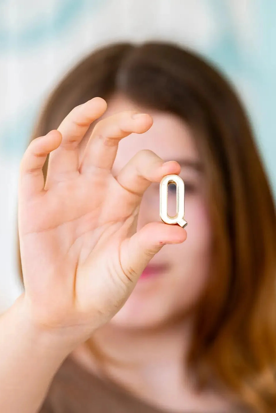 A person holds up a shiny gold letter Q. It is a small, decorative charm, possibly for a necklace or bracelet.