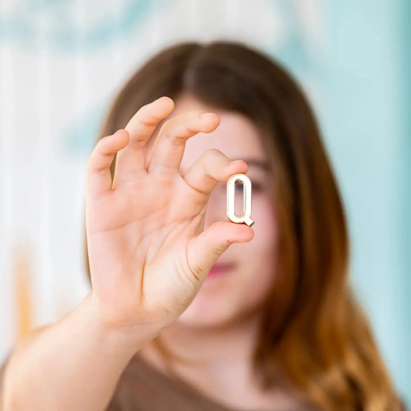 A person holds up a shiny gold letter Q. It is a small, decorative charm, possibly for a necklace or bracelet.