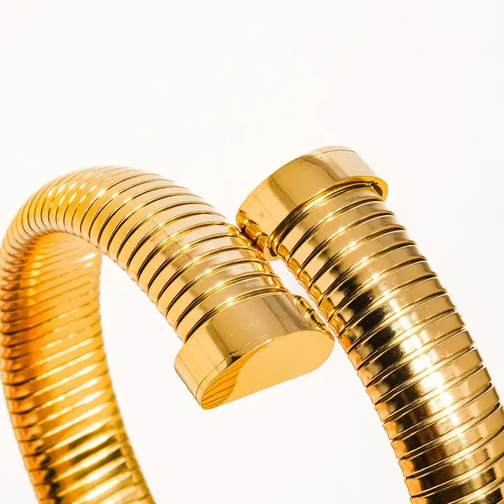 Close-up of a glossy, gold-tone bangle with a flexible, ribbed design and thick end caps. The bangle has a bold, modern