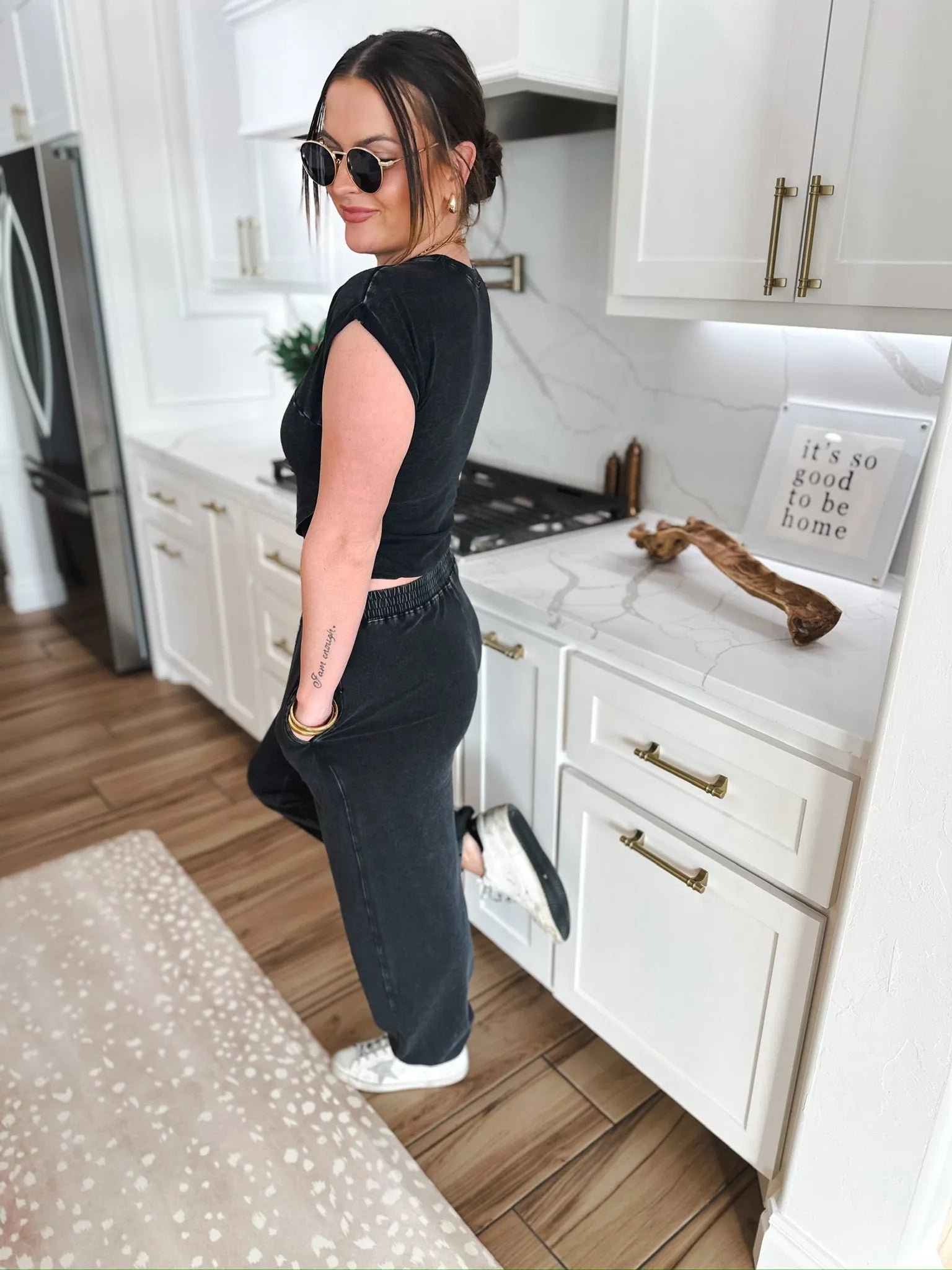 Woman wearing a black Double Zero lounge set, black t-shirt, and black joggers, with sunglasses and white sneakers.