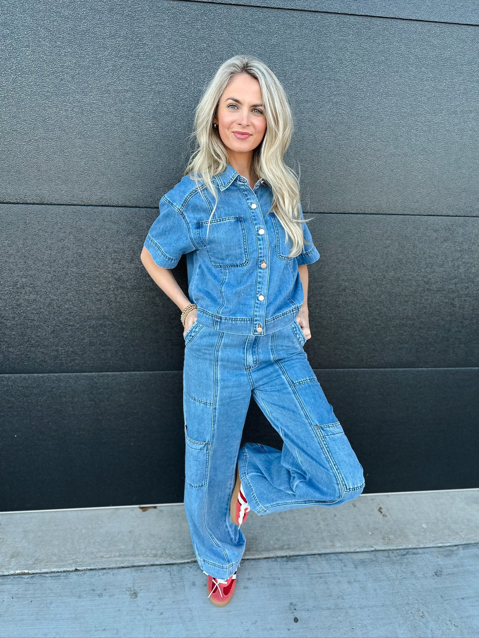 Woman in a light wash denim button-down short-sleeve shirt and matching wide-leg cargo pants, leaning against a dark wall