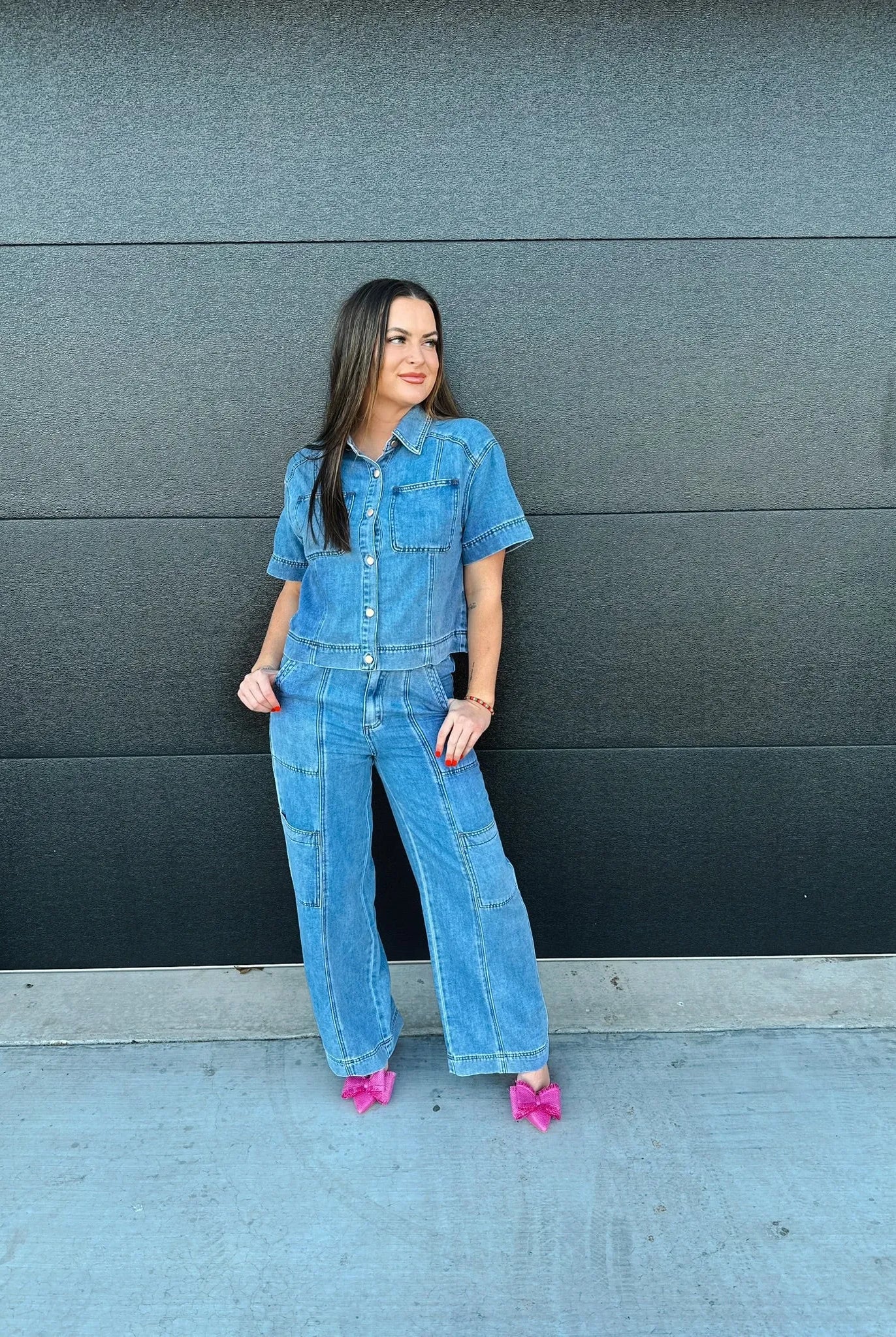 Woman modeling a blue denim button-down shirt and matching cargo pants, paired with pink bow heels, against a dark gray wall.