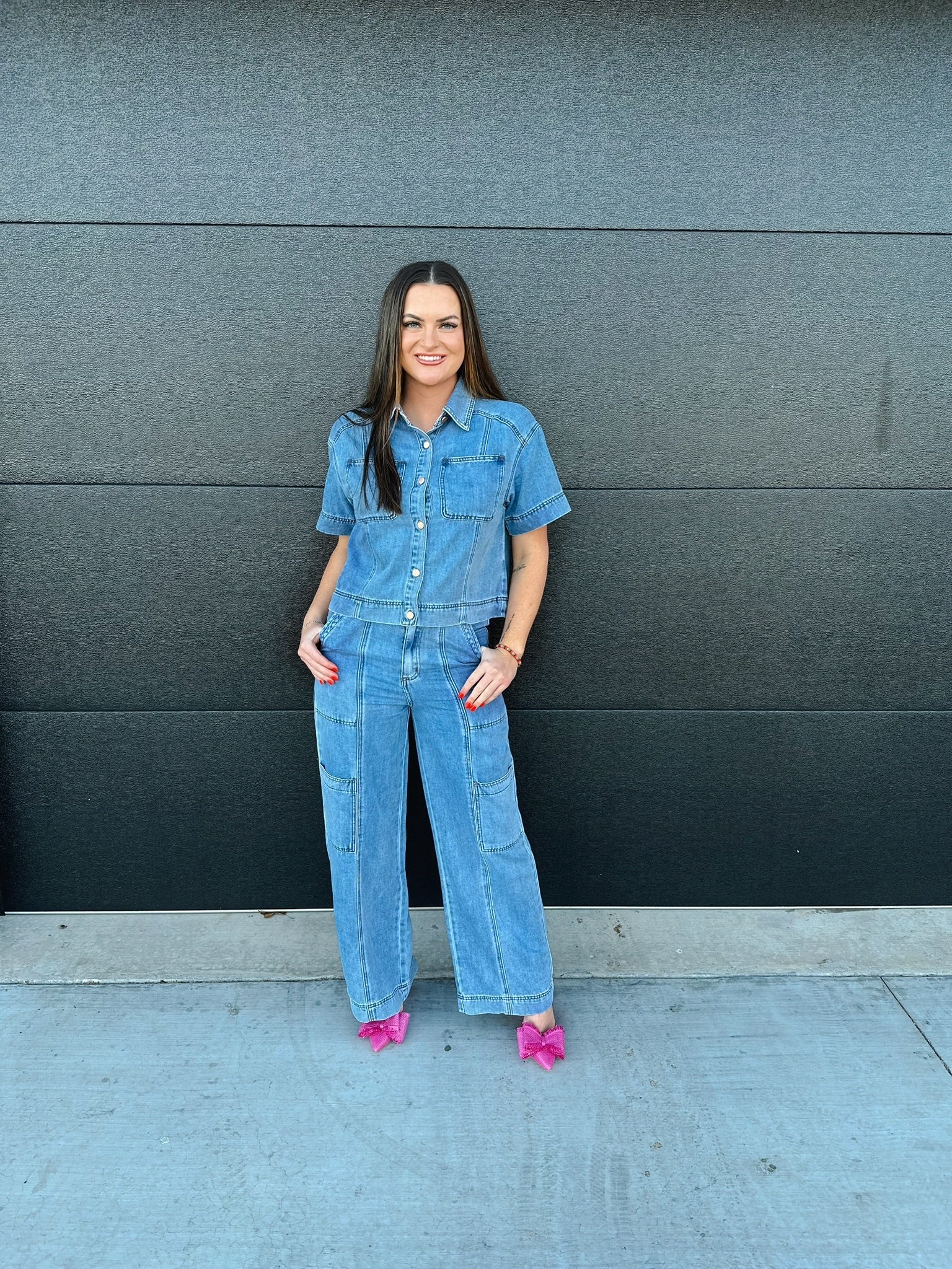 Woman in light wash denim button-down shirt and matching cargo pants. She wears pink bow heels and stands against a dark