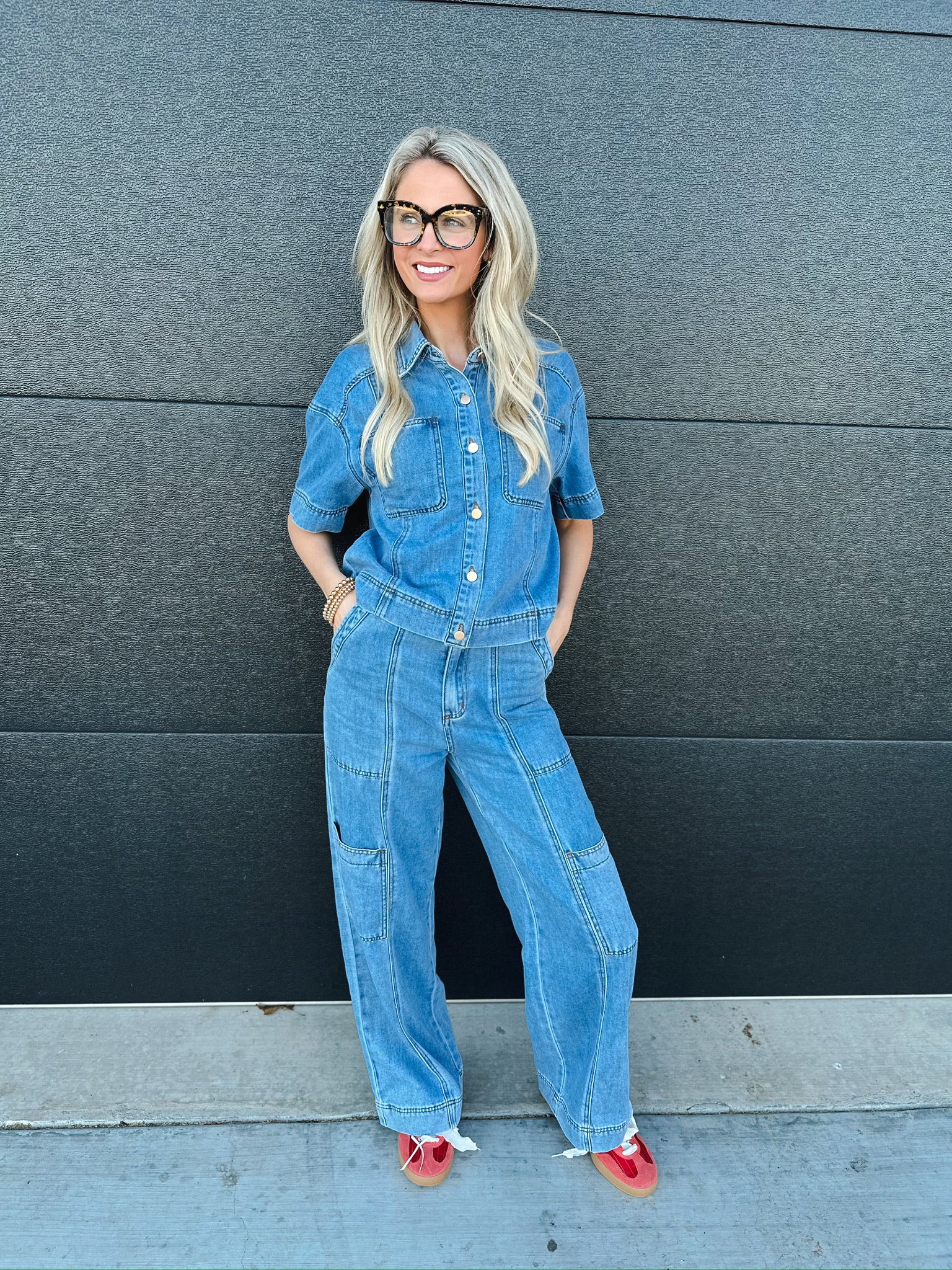 Woman wearing a denim button-down shirt and matching wide-leg cargo jeans. She has on large glasses and red shoes.