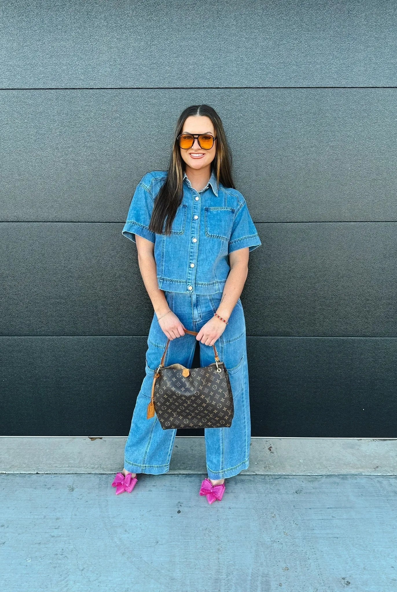 Woman wearing a denim button-down shirt and wide-leg cargo pants, holding a monogrammed brown handbag and pink bow heels.