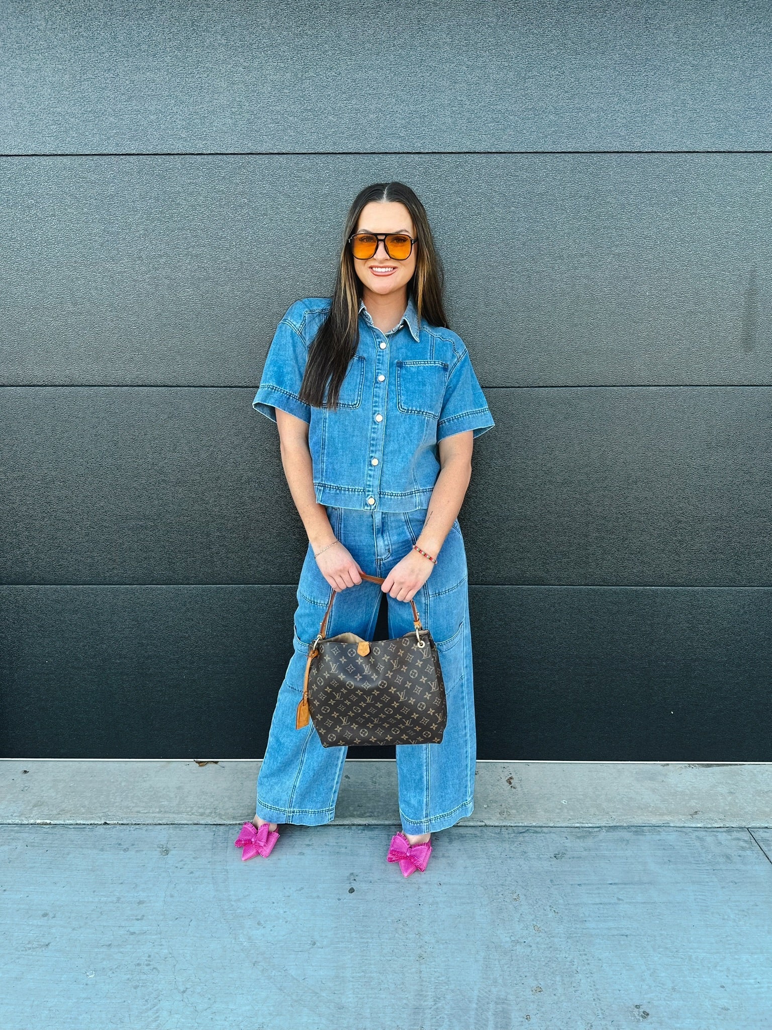 Woman wearing a denim button-down shirt and wide-leg cargo pants, holding a monogrammed brown handbag and pink bow heels.