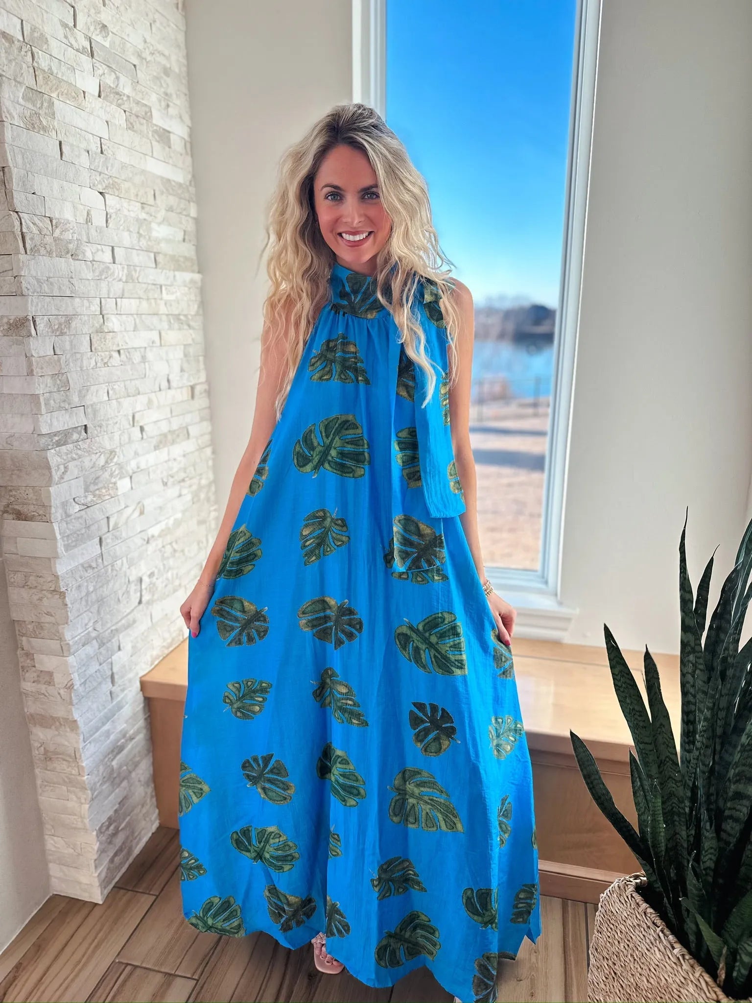 Entro blue maxi dress with tie neck, adorned with a green palm leaf print. Model is smiling, standing indoors by a window.
