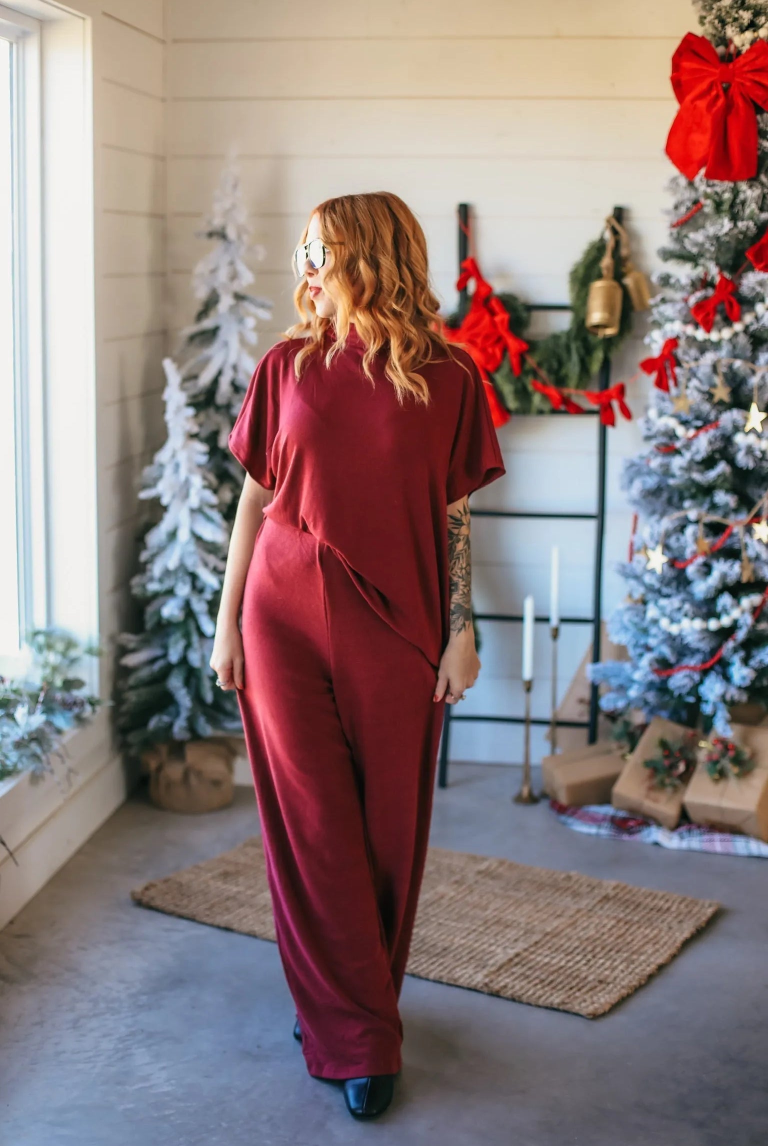 Woman wearing a burgundy ribbed jumpsuit with wide legs, looking out a window next to a decorated Christmas tree.