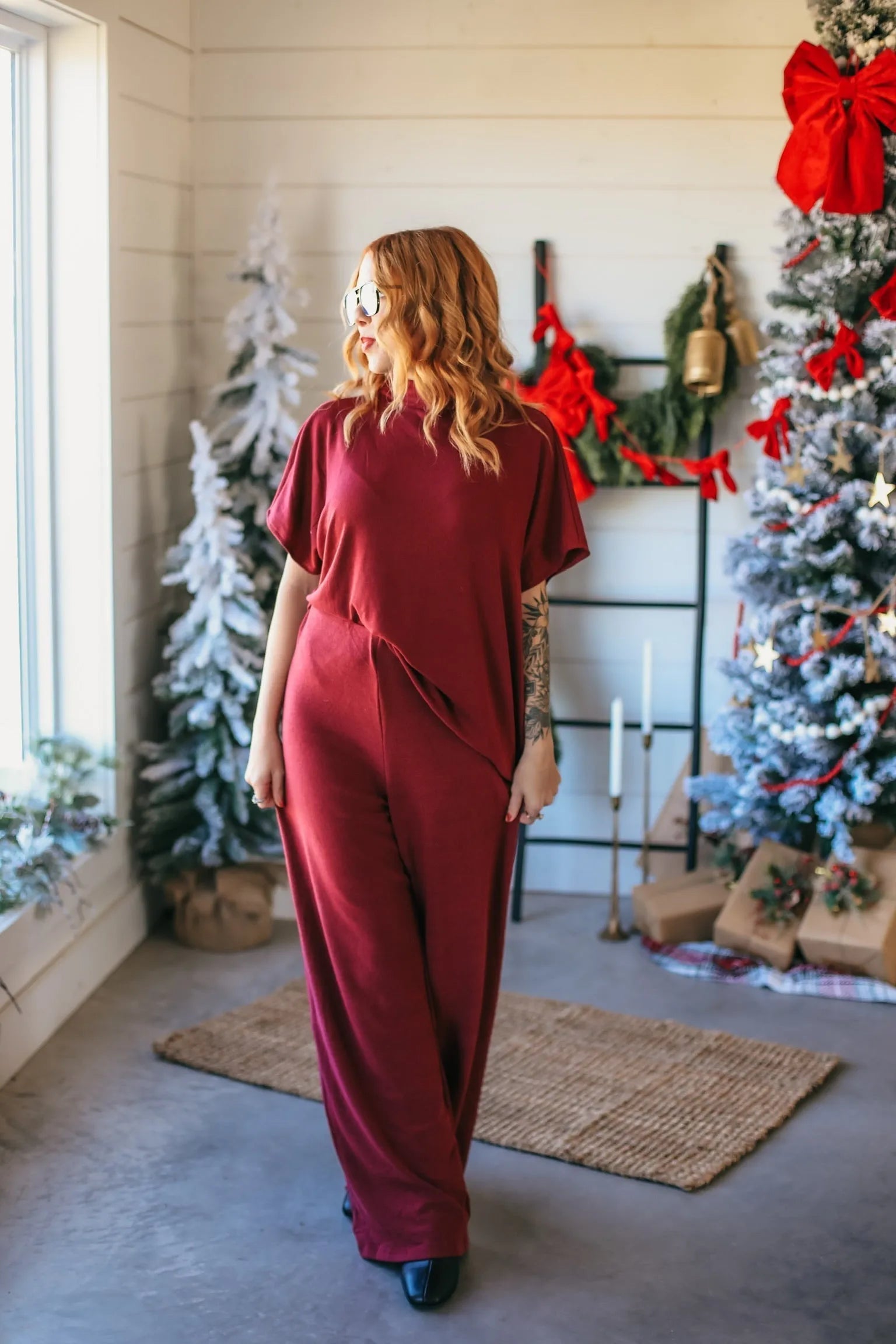 Woman wearing a burgundy ribbed jumpsuit with wide legs, looking out a window next to a decorated Christmas tree.