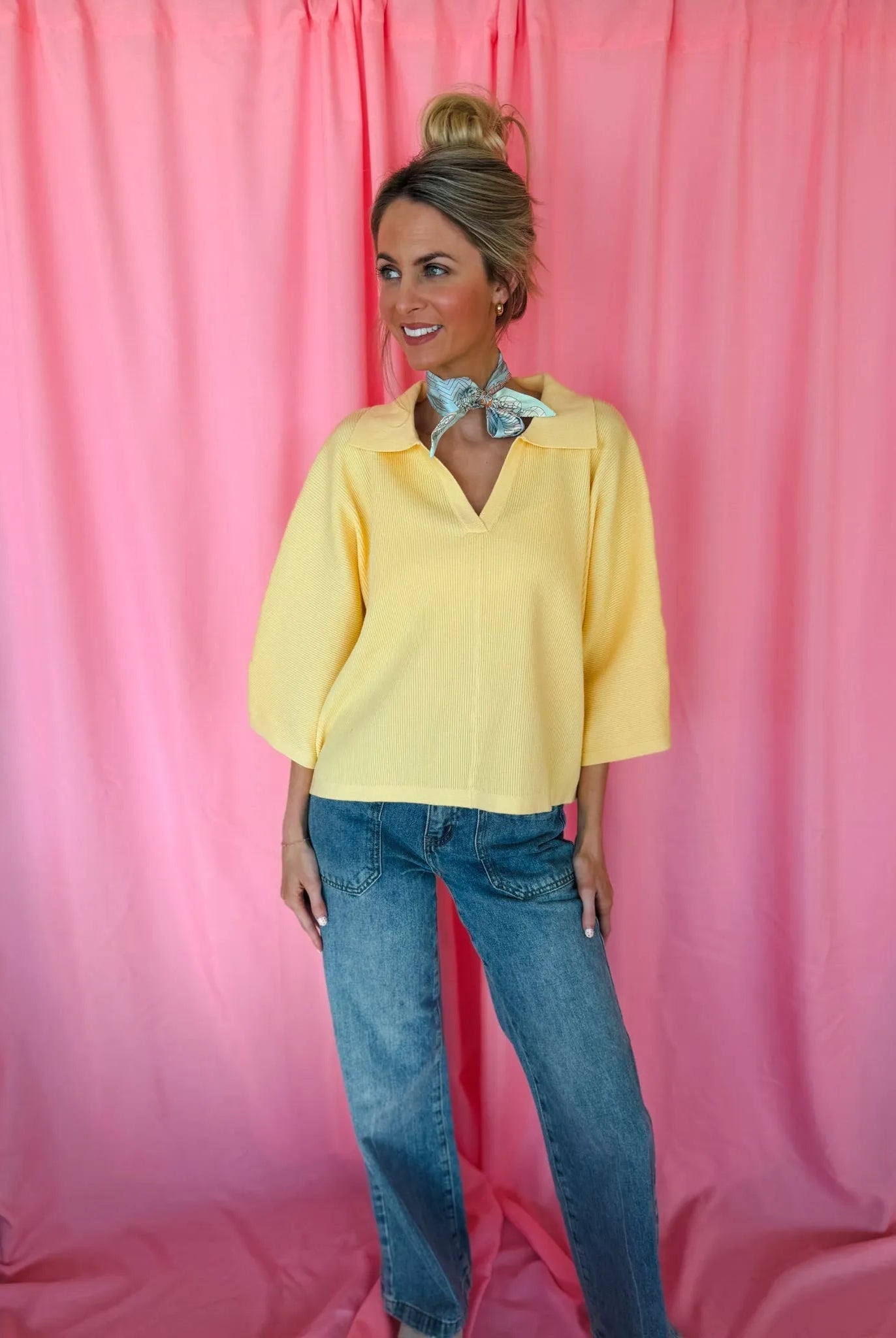 A woman wears a butter yellow ribbed sweater top with a V-neck and collar, paired with blue denim jeans. A light blue