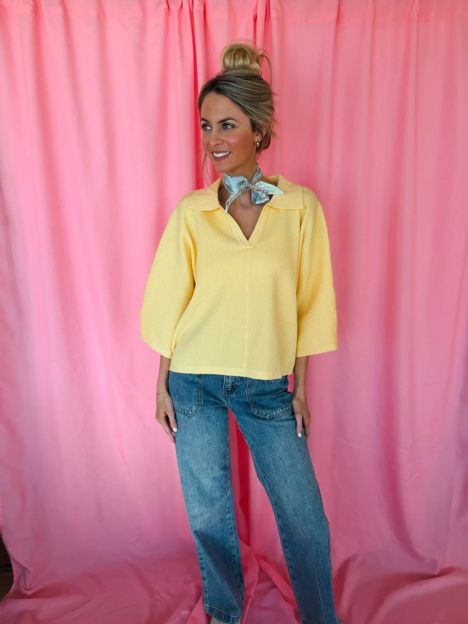 A woman wears a butter yellow ribbed sweater top with a V-neck and collar, paired with blue denim jeans. A light blue