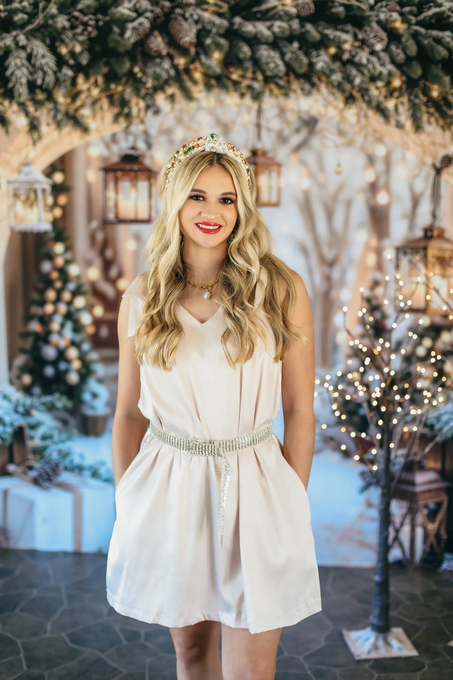Woman in champagne V-neck cocktail dress with rhinestone belt. Festive background with Christmas trees and lights.