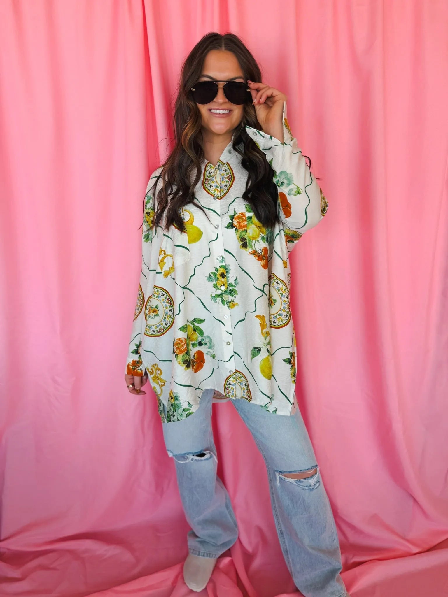 Woman wearing an oversized white button-down top with a vibrant citrus and floral print. She also wears ripped light-wash