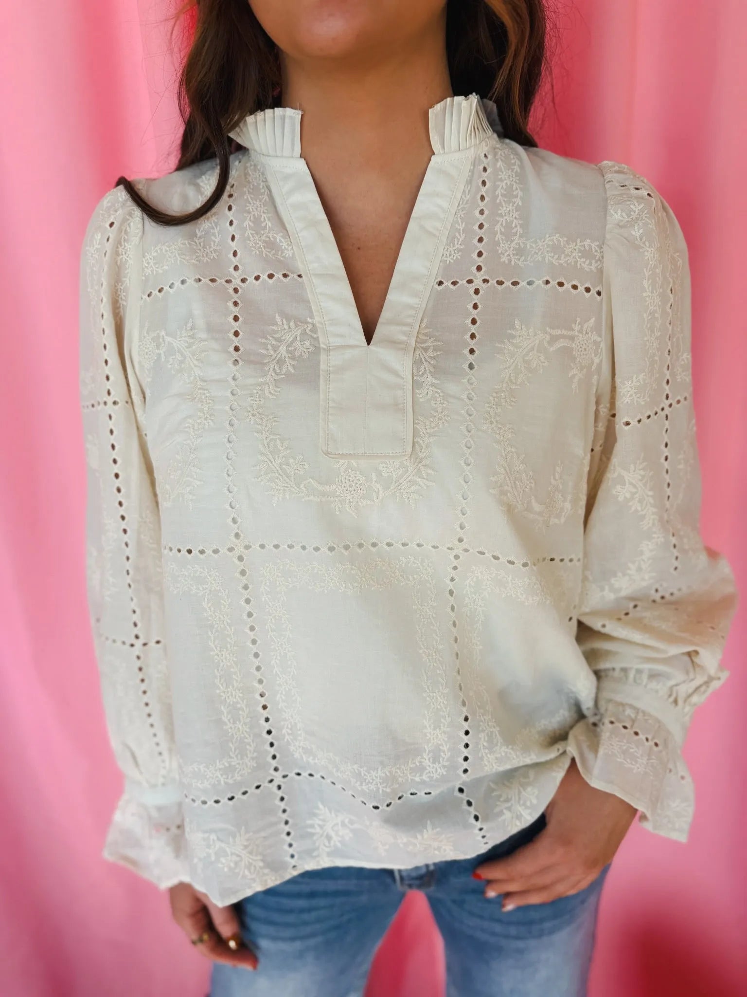 Cream eyelet long sleeve blouse with embroidered floral details and a v-neck collar, set against a pink backdrop.