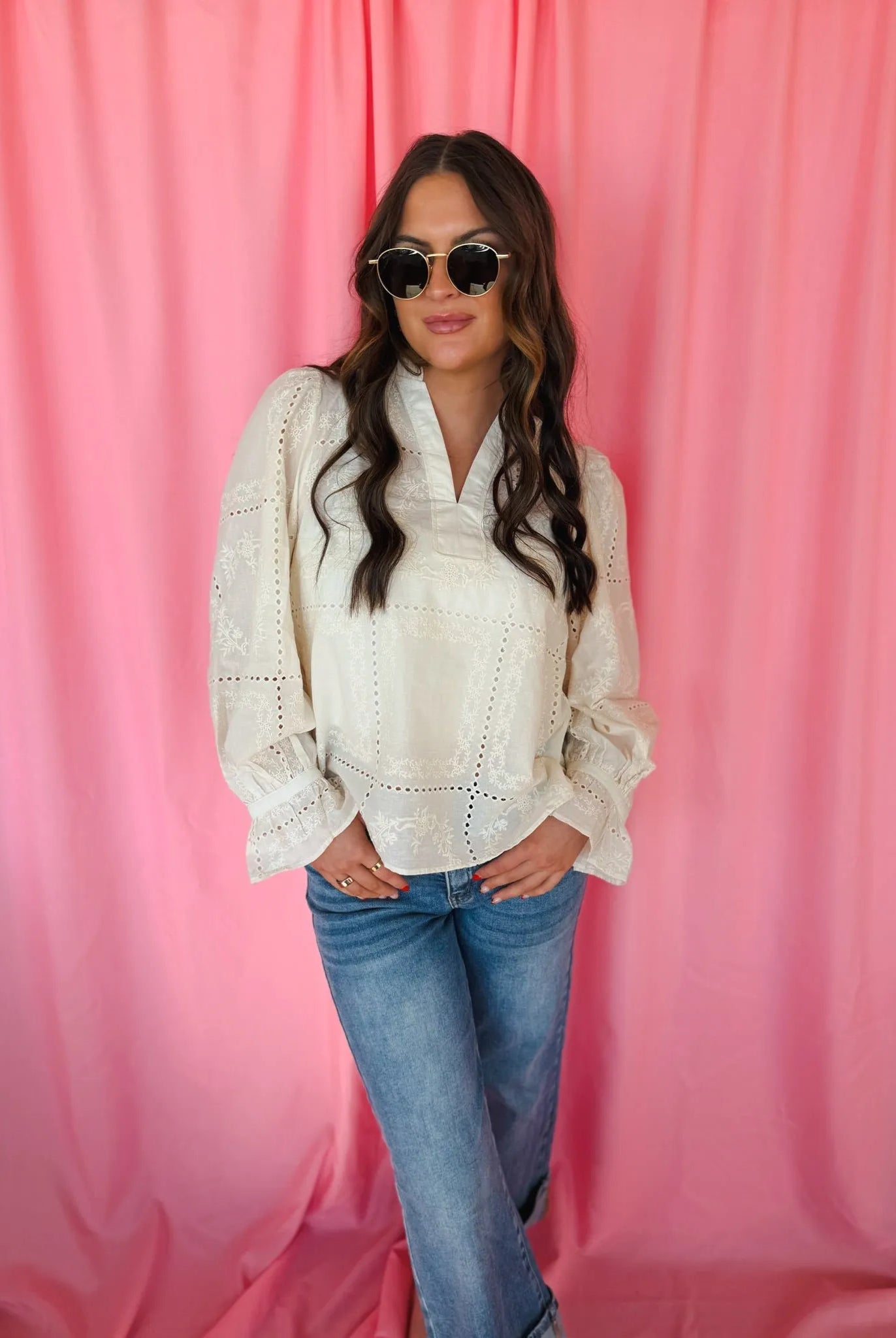 Woman wearing a cream eyelet long sleeve blouse with a v-neck and bell sleeves, paired with blue jeans. She has on