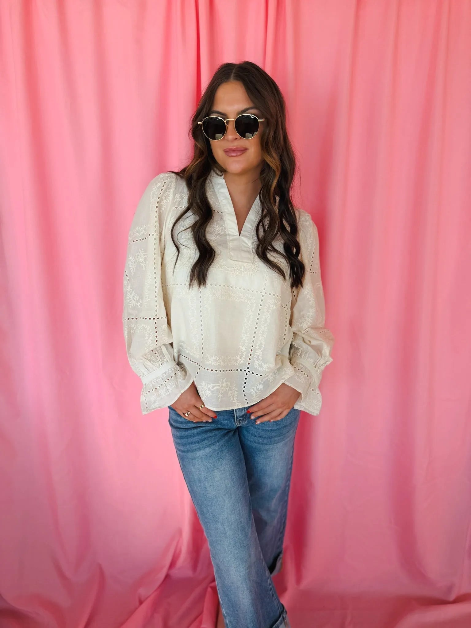 Woman wearing a cream eyelet long sleeve blouse with a v-neck and bell sleeves, paired with blue jeans. She has on