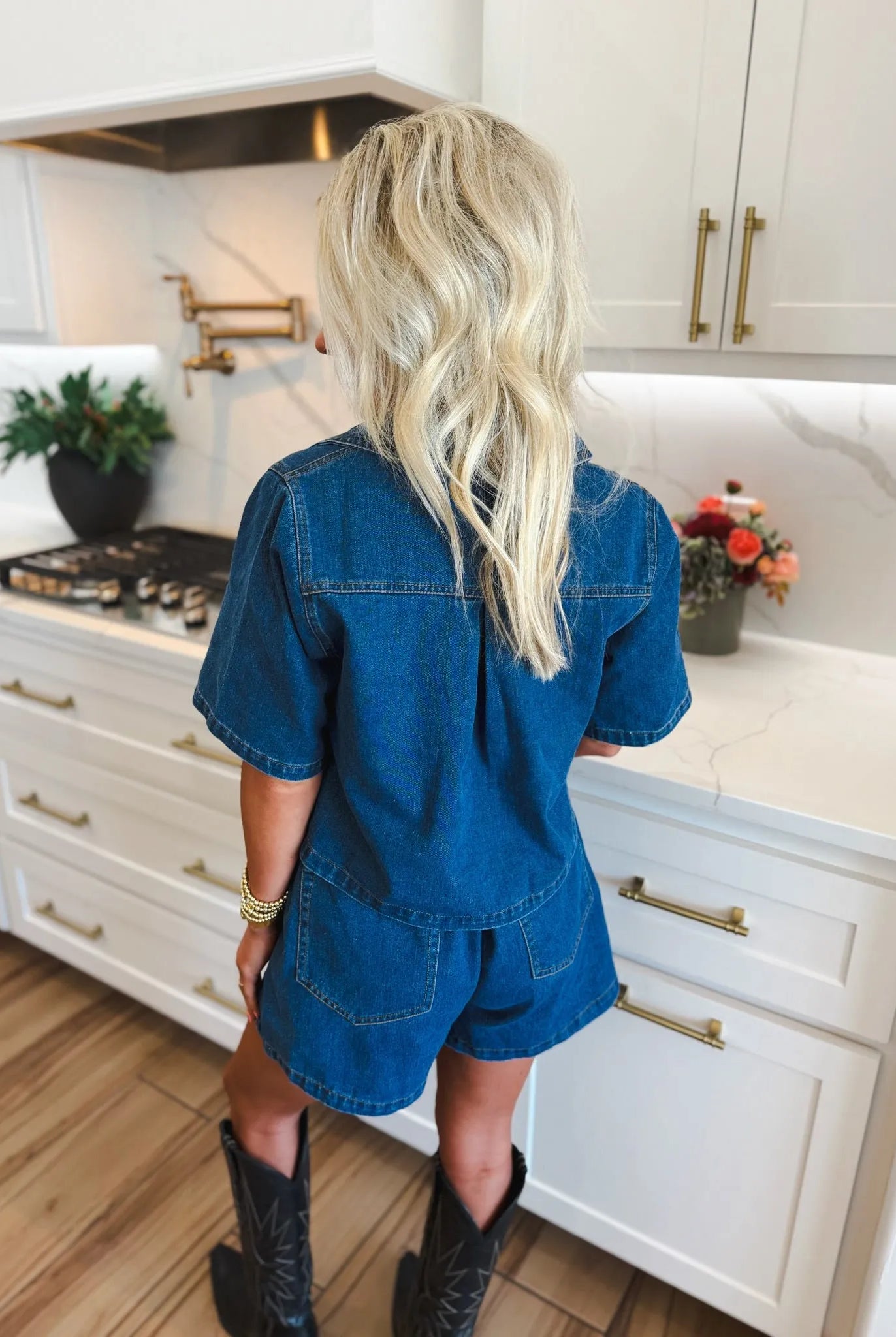 Woman from behind wearing a dark denim skort set with short sleeves and shorts. She has blonde hair and black cowboy boots