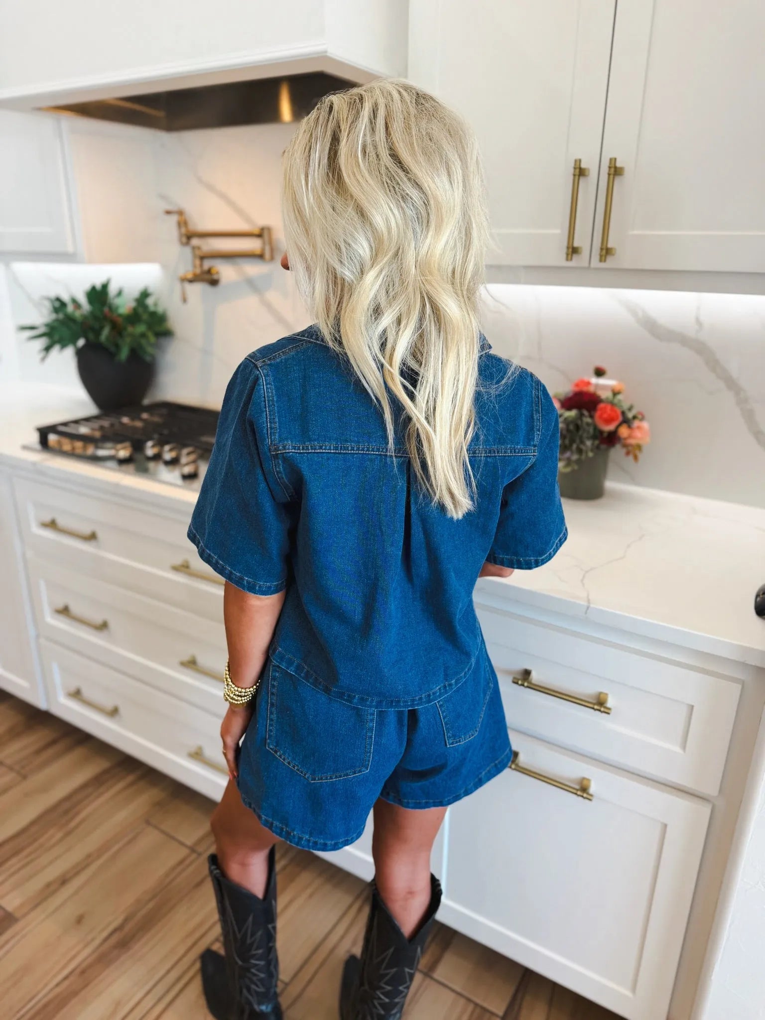 Woman from behind wearing a dark denim skort set with short sleeves and shorts. She has blonde hair and black cowboy boots