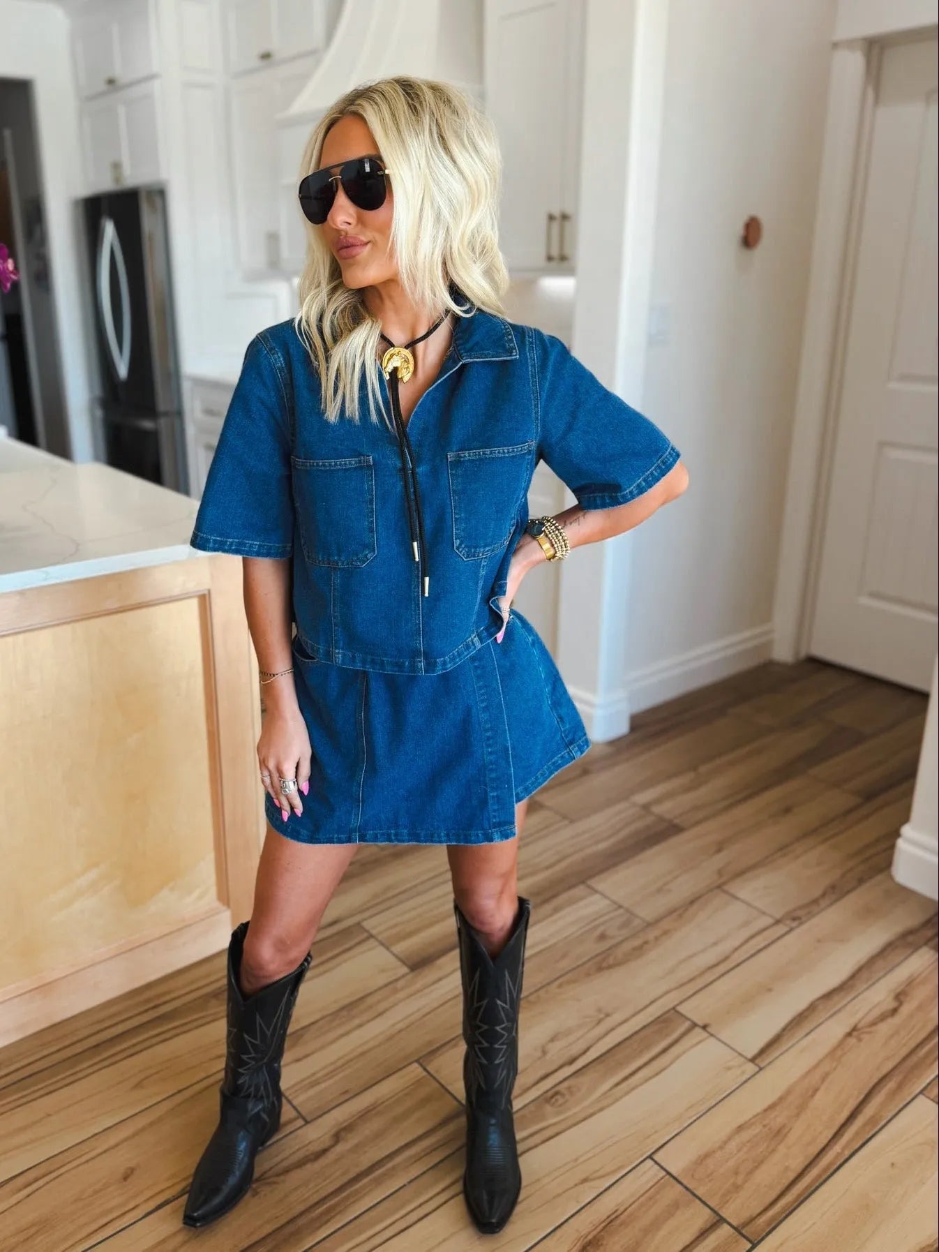 Woman wearing a dark denim skort set with short sleeves and two chest pockets, paired with black cowboy boots and sunglasses.