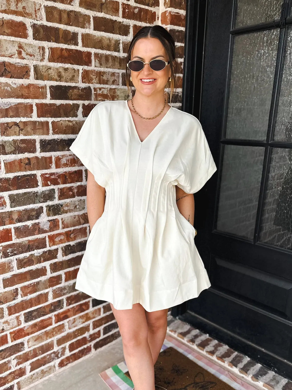 Woman wearing a cream V-neck dress with pleats and pockets, featuring short flutter sleeves and a flared skirt.