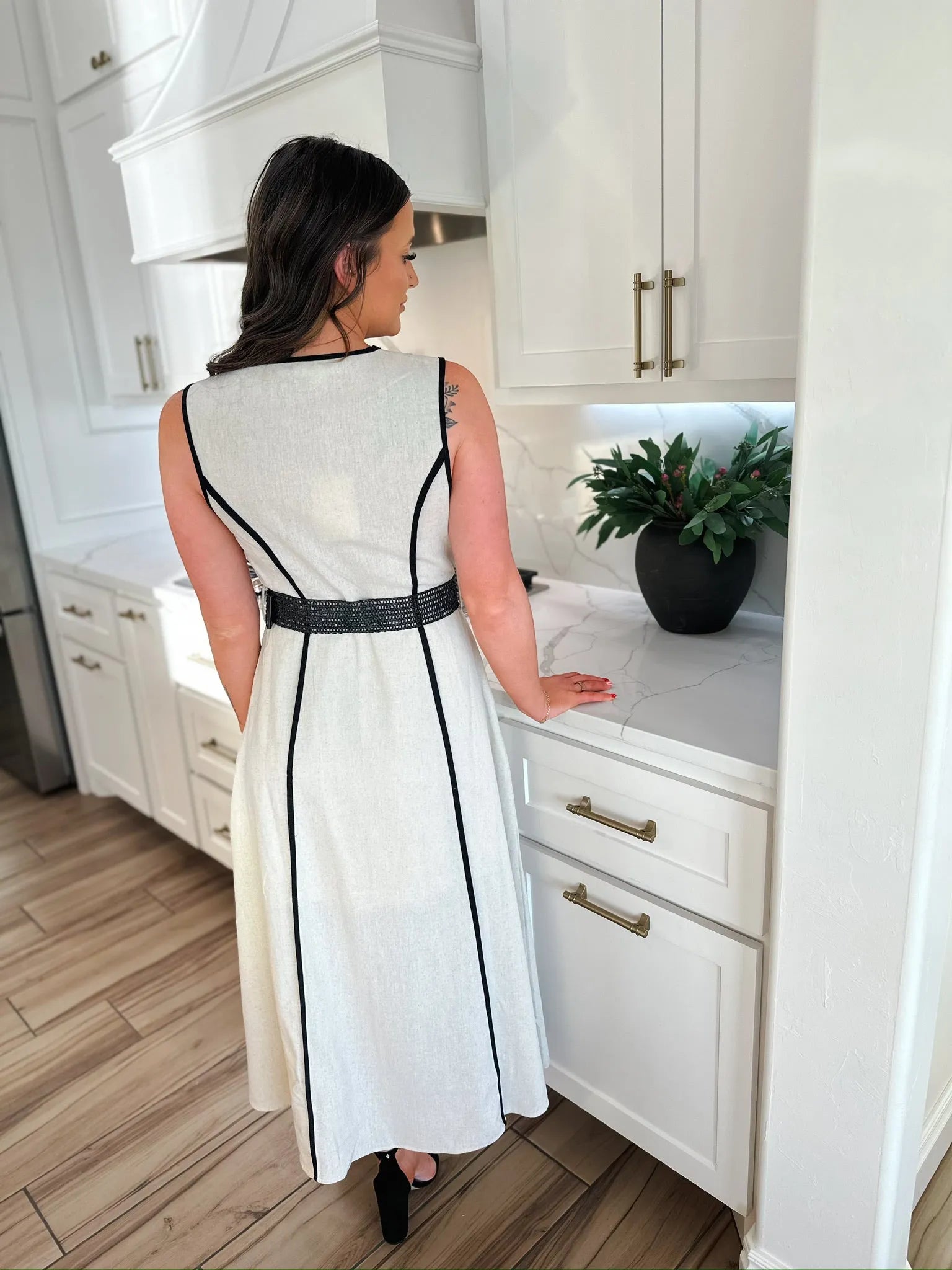 Woman in an ecru linen blend midi dress with black trim and a black belt, standing in a modern kitchen.