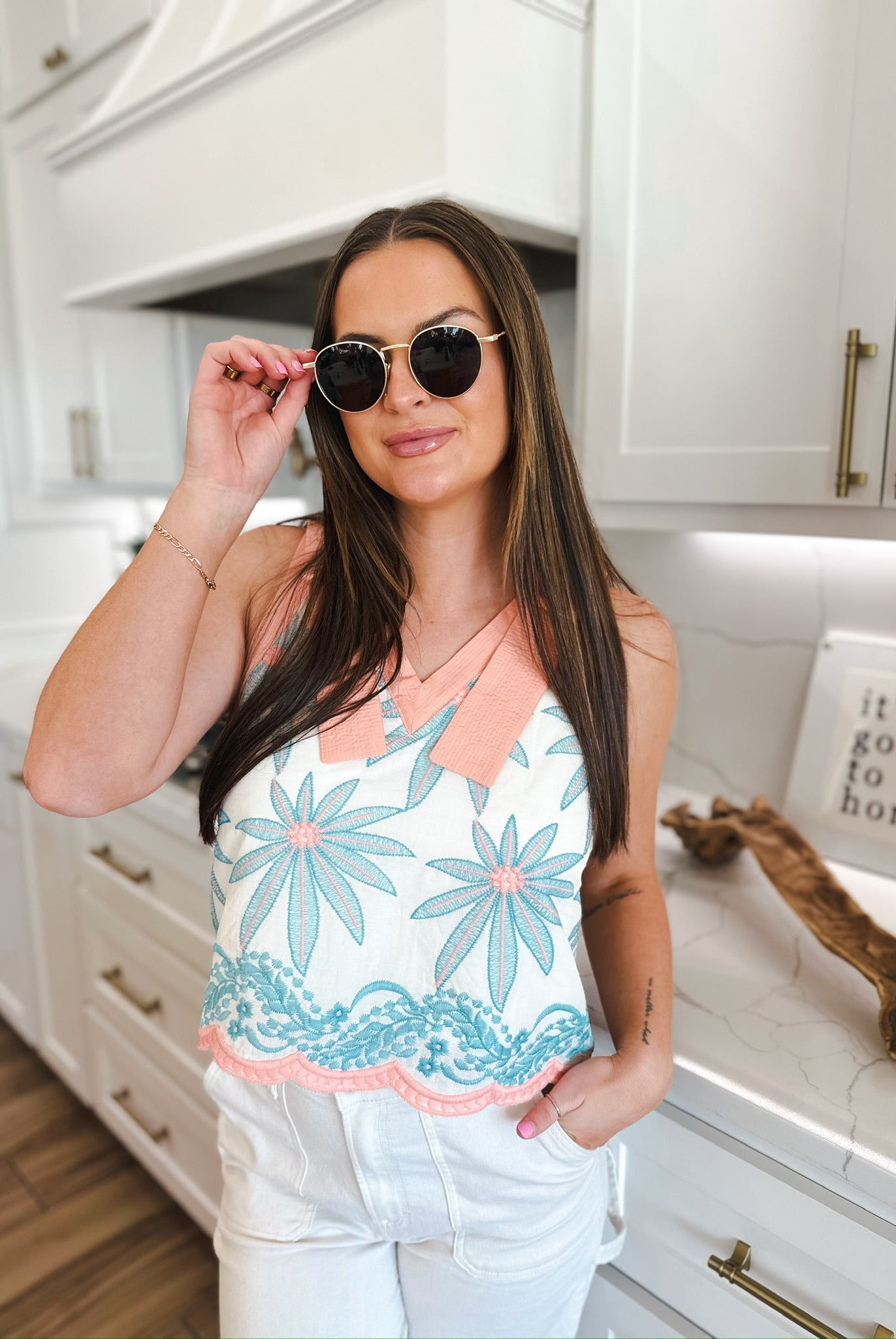 Entro white sleeveless blouse with blue floral embroidery and pink trim. Model wears sunglasses and white pants.