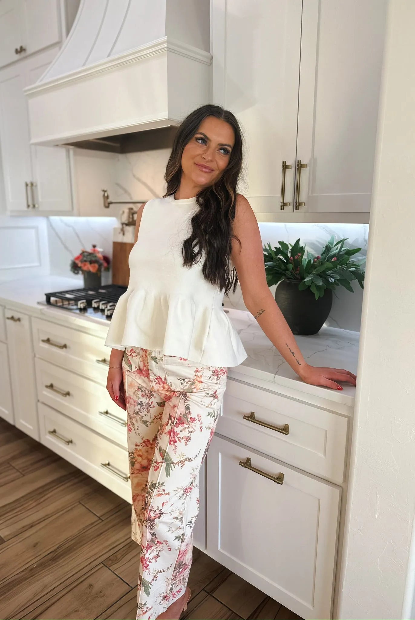 Entro Ivory sleeveless peplum top paired with floral print pants. Woman standing in a modern kitchen.