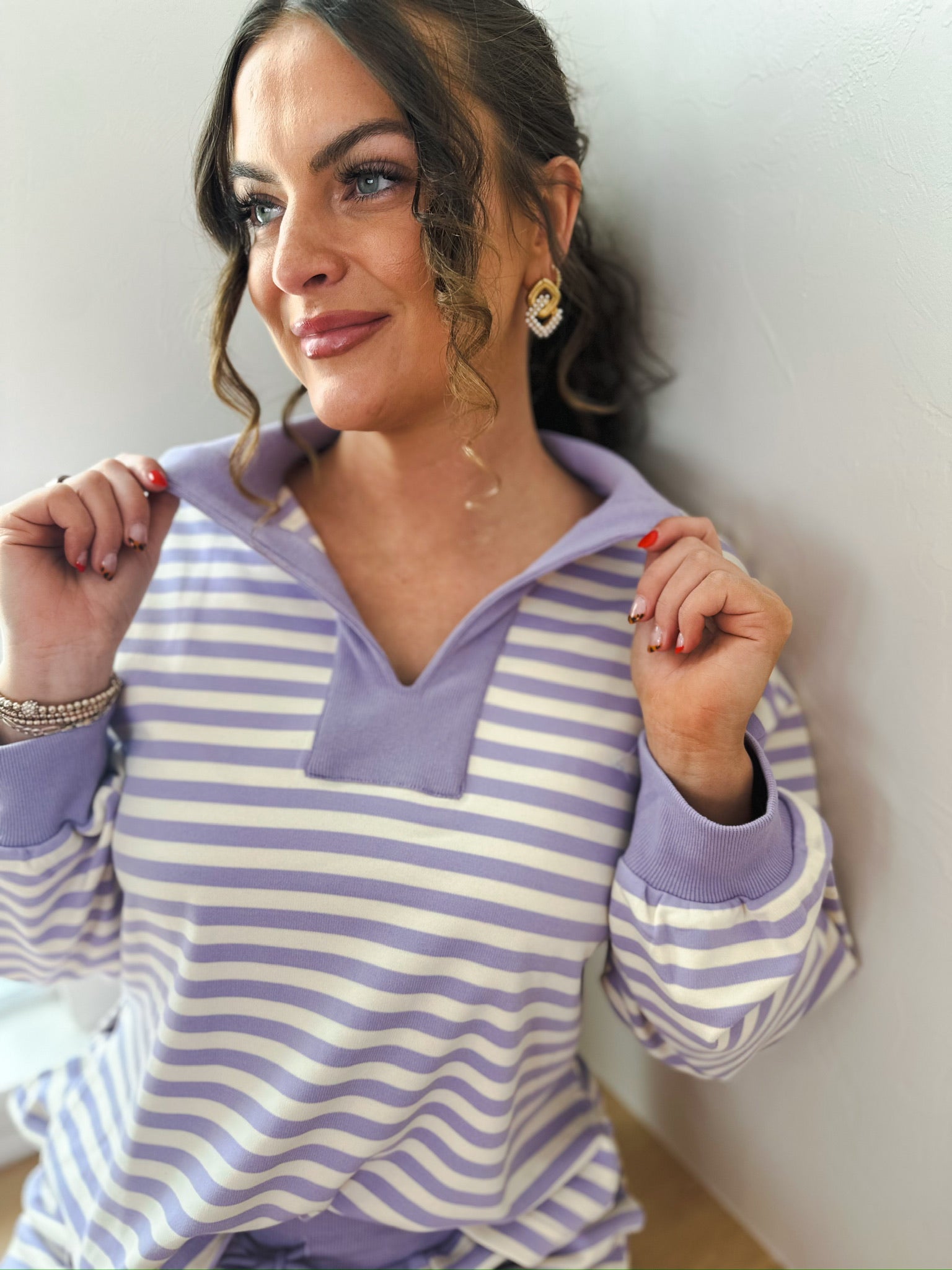 Woman in a lavender and cream striped pullover with a collar. She wears gold earrings and a bracelet.