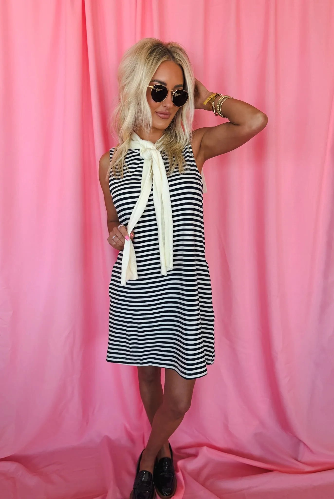 Blonde woman in Entro navy & cream striped sleeveless dress, featuring a tied neck scarf. Wearing sunglasses & loafers