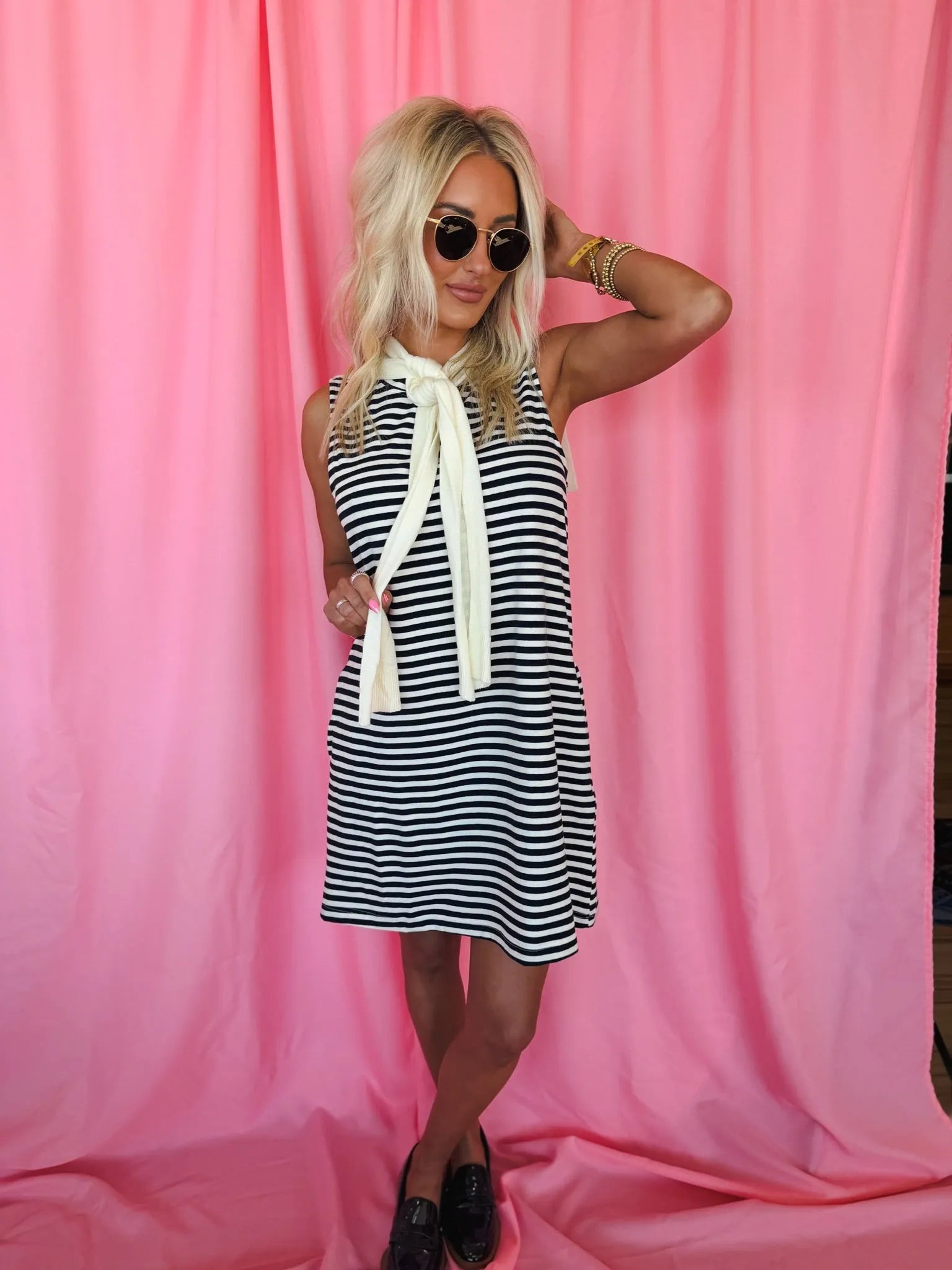 Blonde woman in Entro navy & cream striped sleeveless dress, featuring a tied neck scarf. Wearing sunglasses & loafers