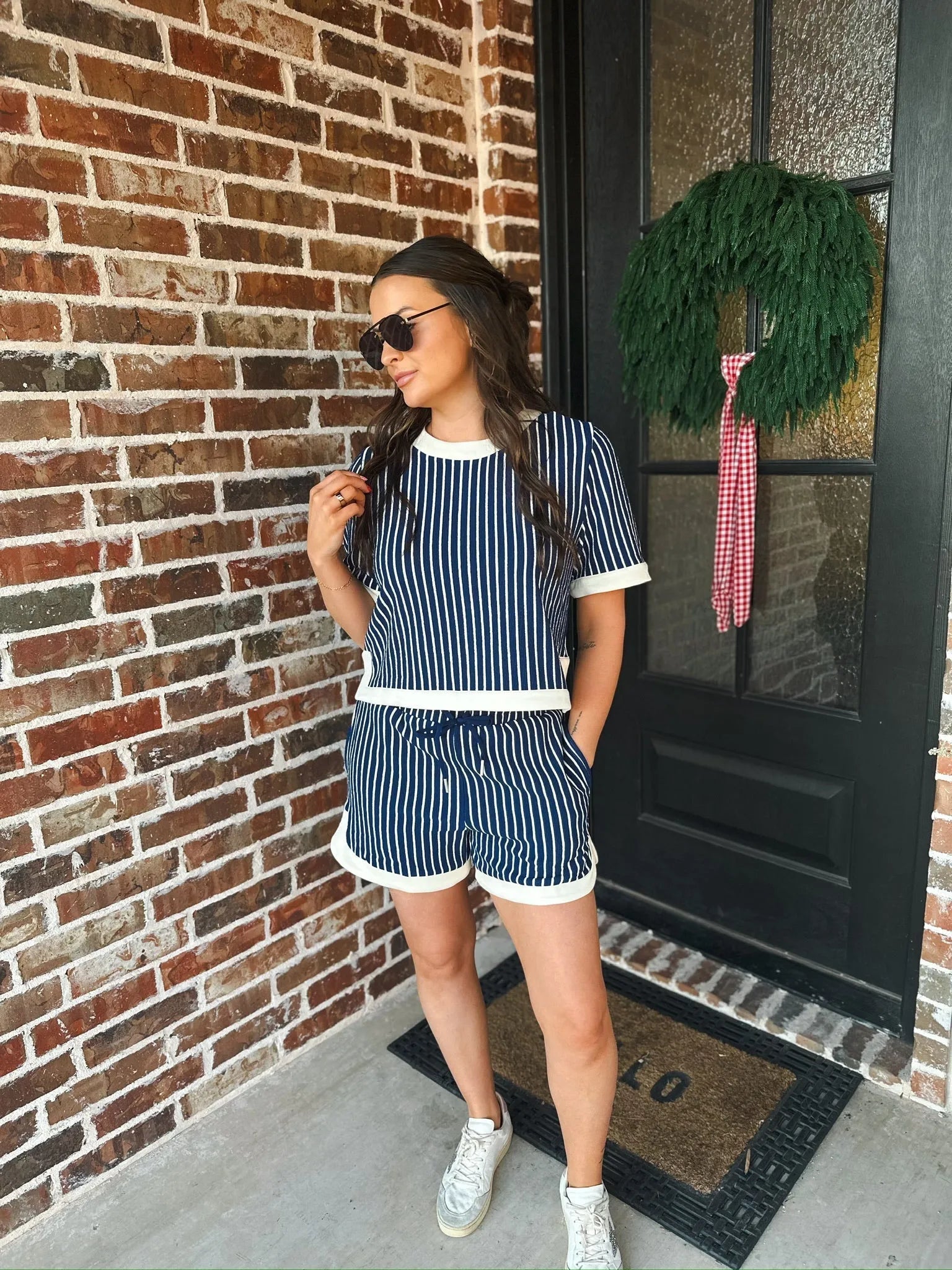 Woman wearing a navy and cream striped two-piece short set with white trim, standing outdoors by a brick wall and a black