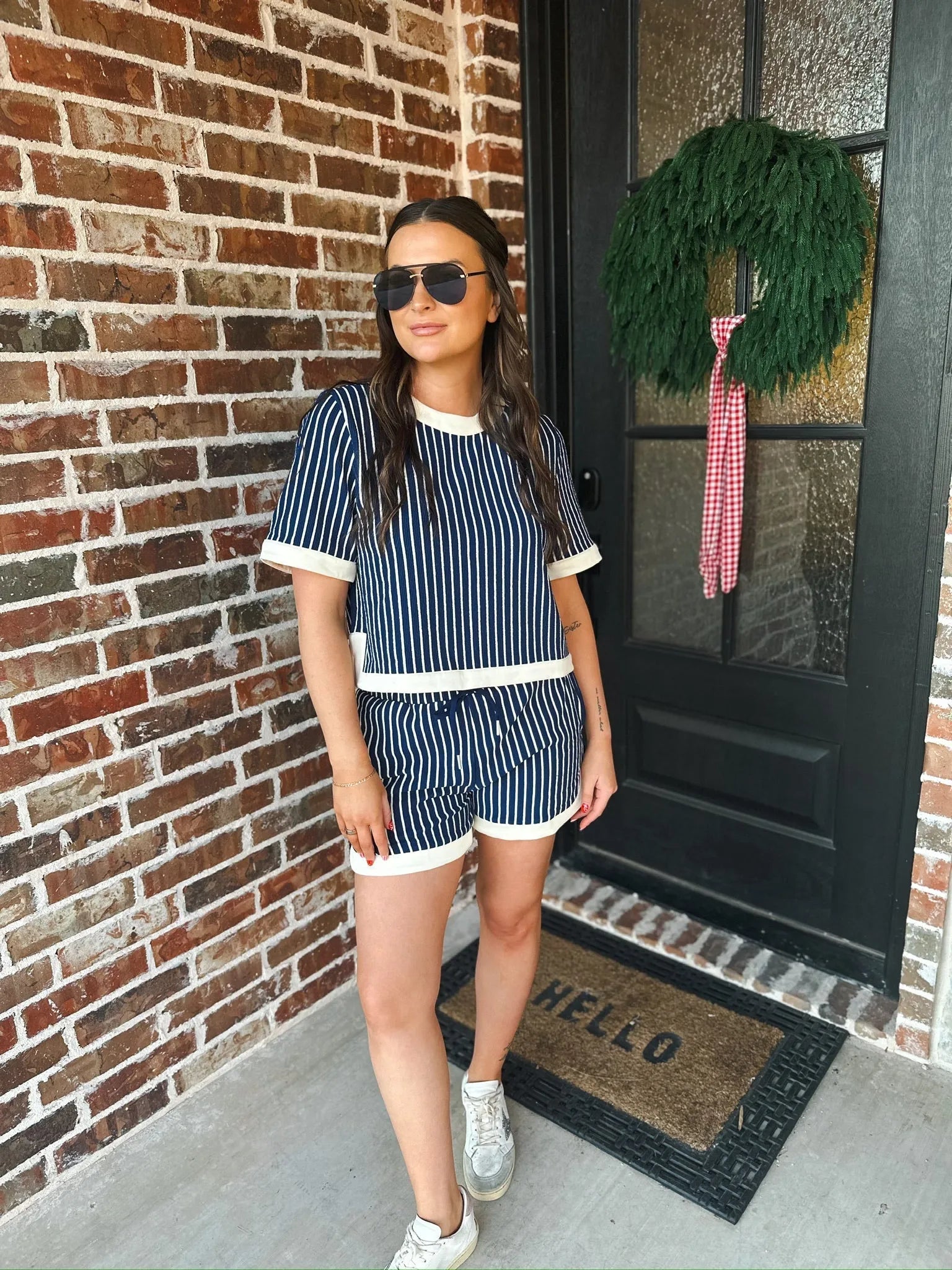 Woman wearing a navy and cream striped two-piece short set with cream trim, standing on a porch.