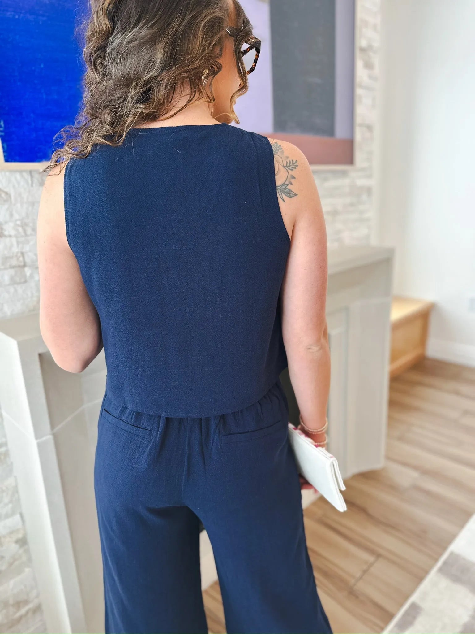Back view of a woman wearing a navy blue sleeveless button-down blouse and navy blue wide-leg pants. She has curly brown