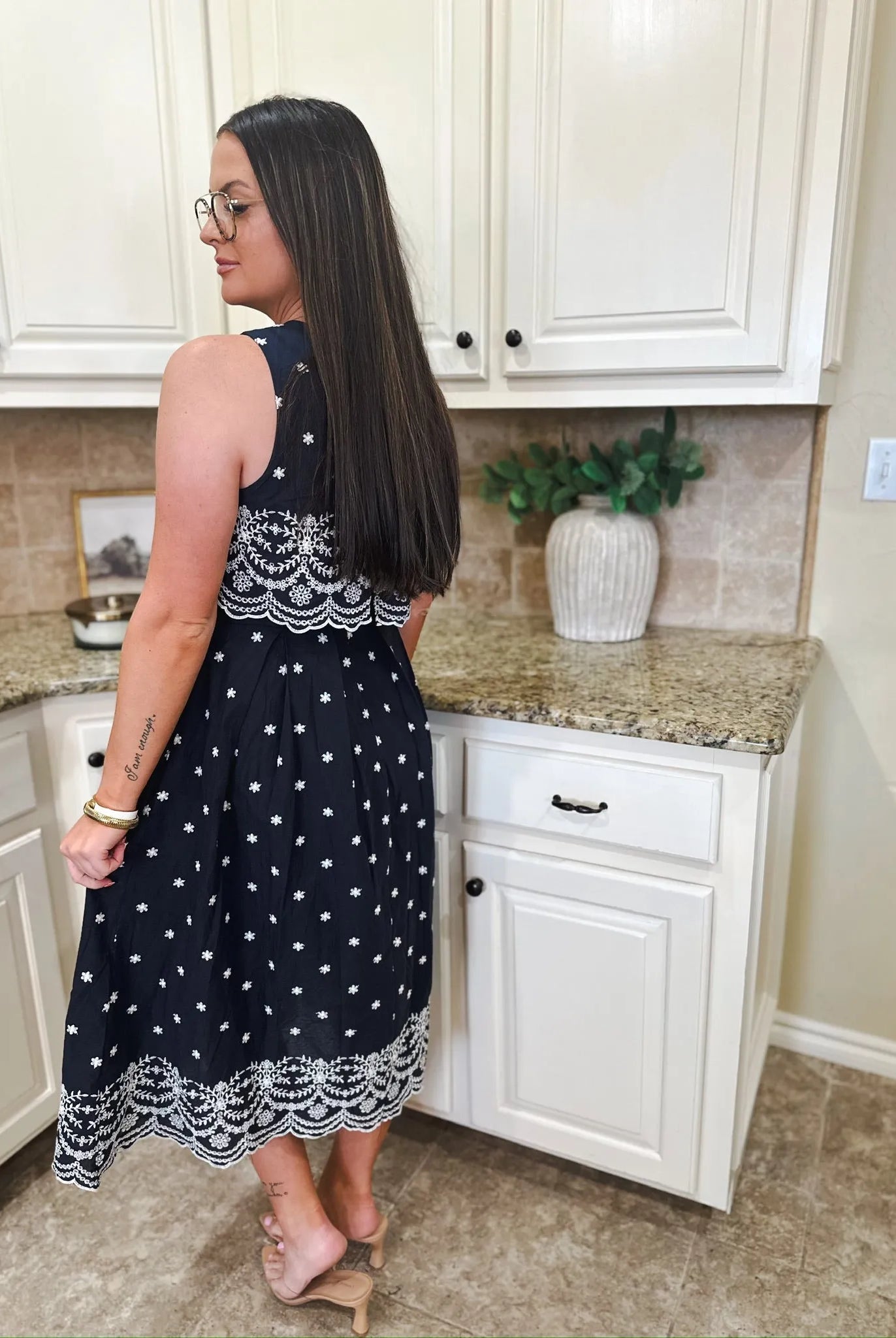 Navy blue sleeveless midi dress with white eyelet trim and white floral embroidery. A woman wears it in a kitchen.