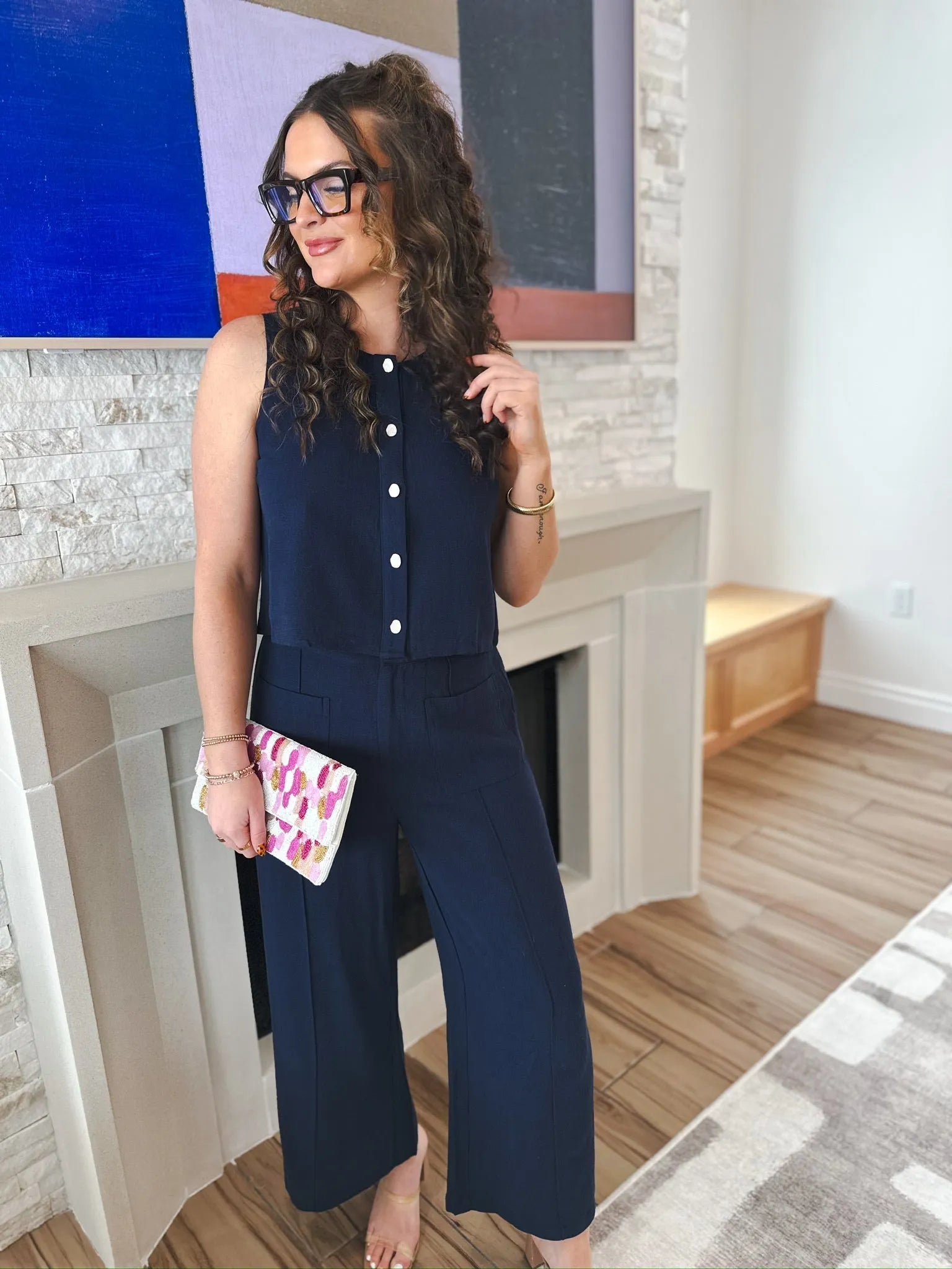 Woman wearing navy blue wide-leg trousers and a matching sleeveless top with white buttons. She holds a colorful clutch and