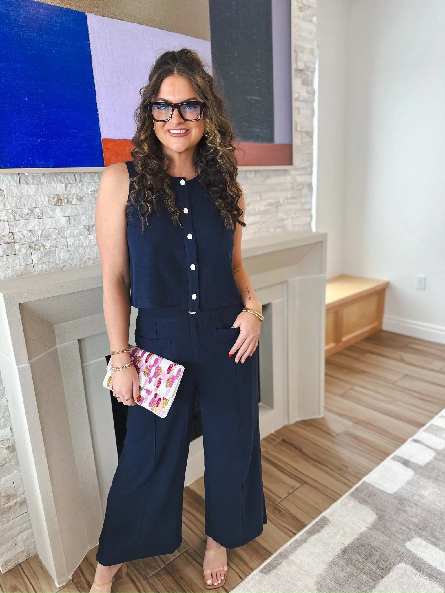 Woman wearing navy culotte trousers and matching sleeveless top with white buttons. She holds a patterned clutch.