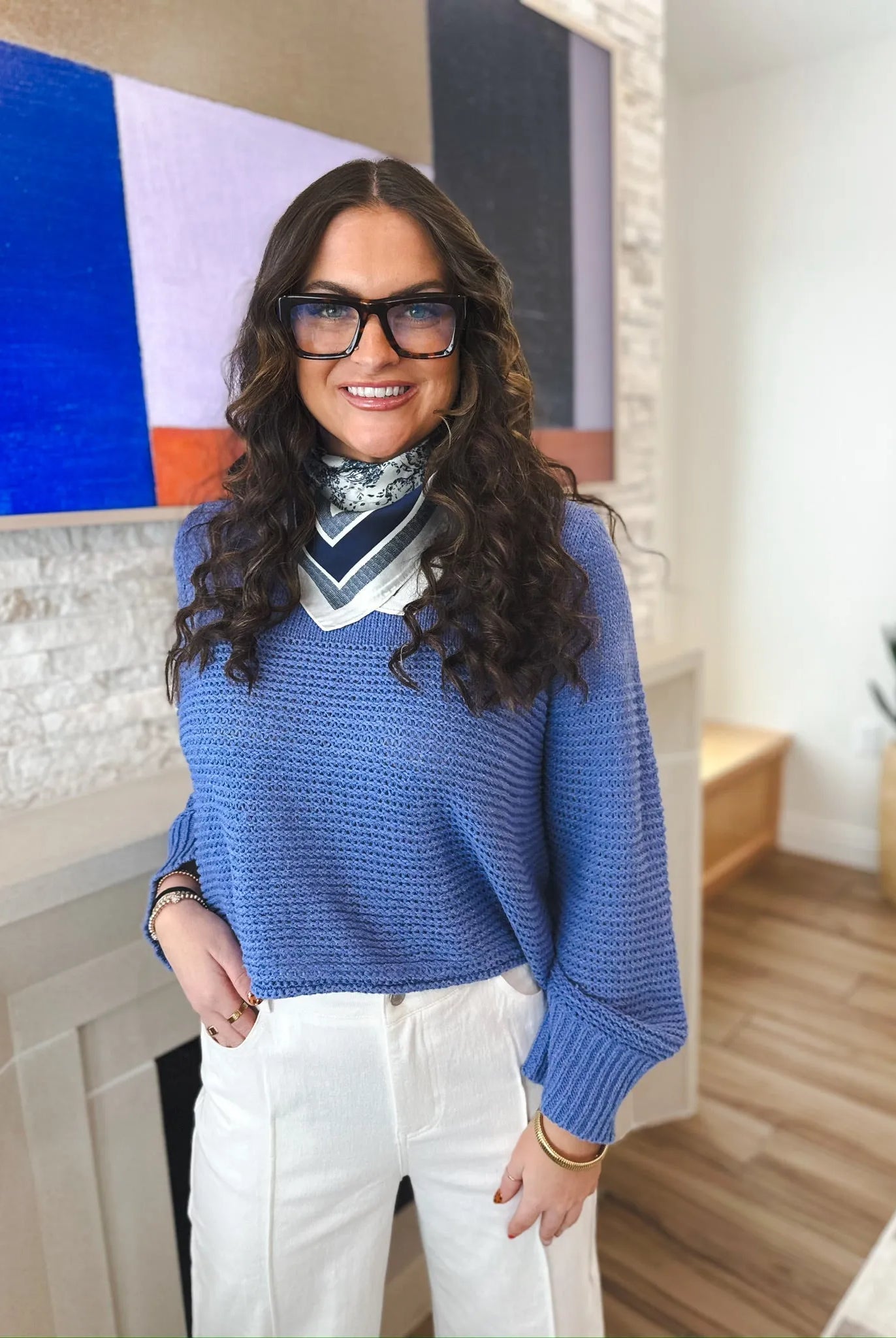 Woman wearing a periwinkle knit sweater with a v-neck and a patterned scarf. She has on white pants and glasses.