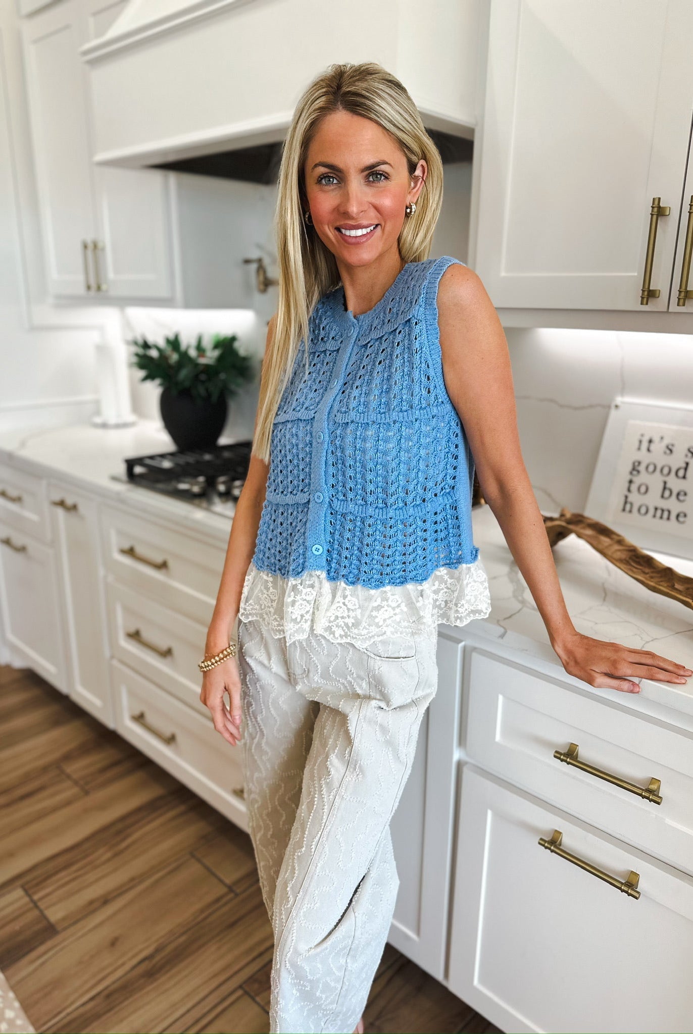 Blue crochet sleeveless cardigan sweater with white lace trim, paired with textured beige pants. Woman standing in a kitchen.