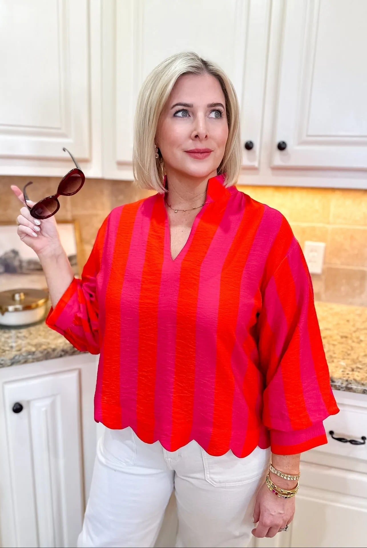 Woman wearing a pink and red striped 3/4 sleeve blouse with a scalloped hem and white pants.