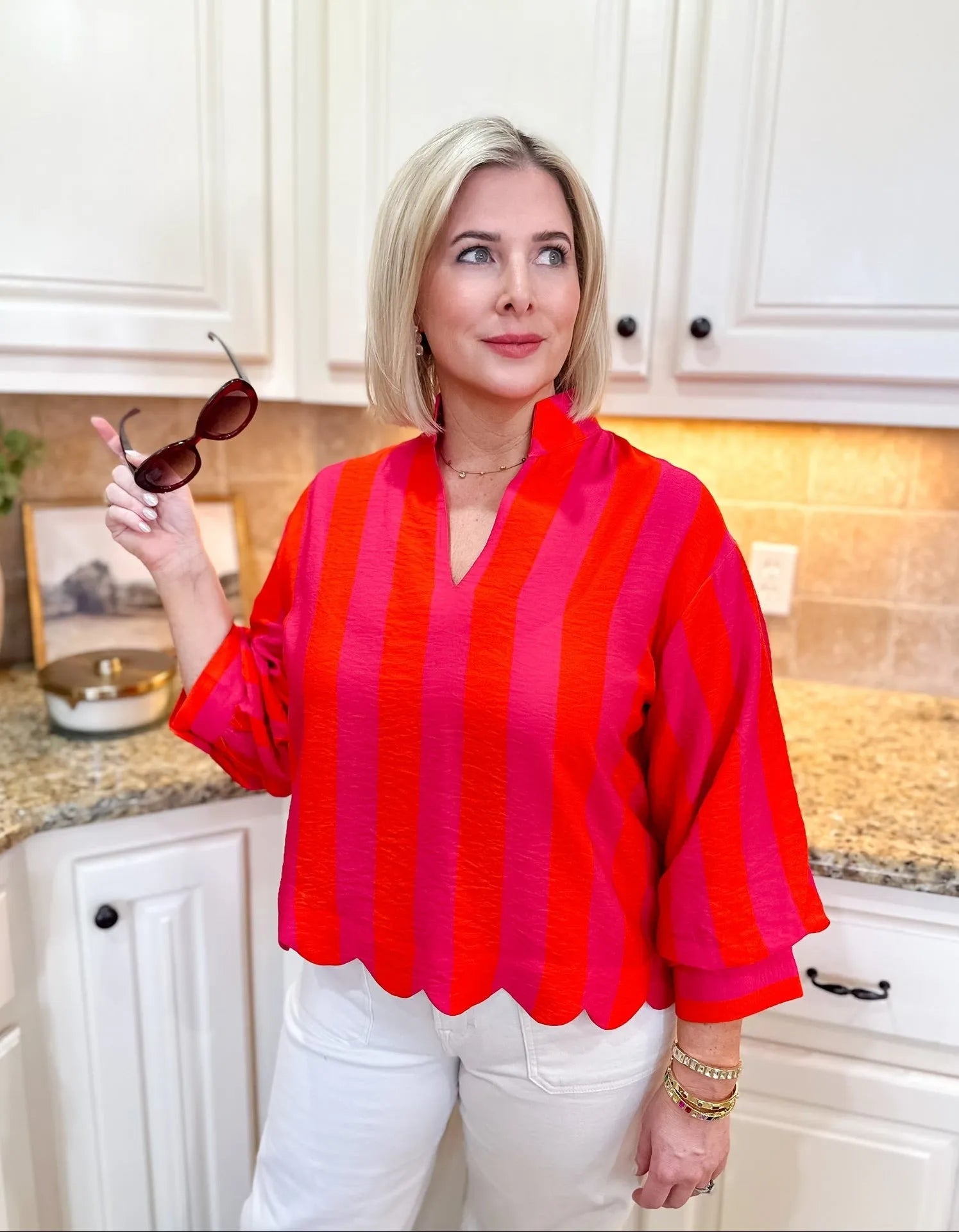 Woman wearing a pink and red striped 3/4 sleeve blouse with a scalloped hem and white pants.