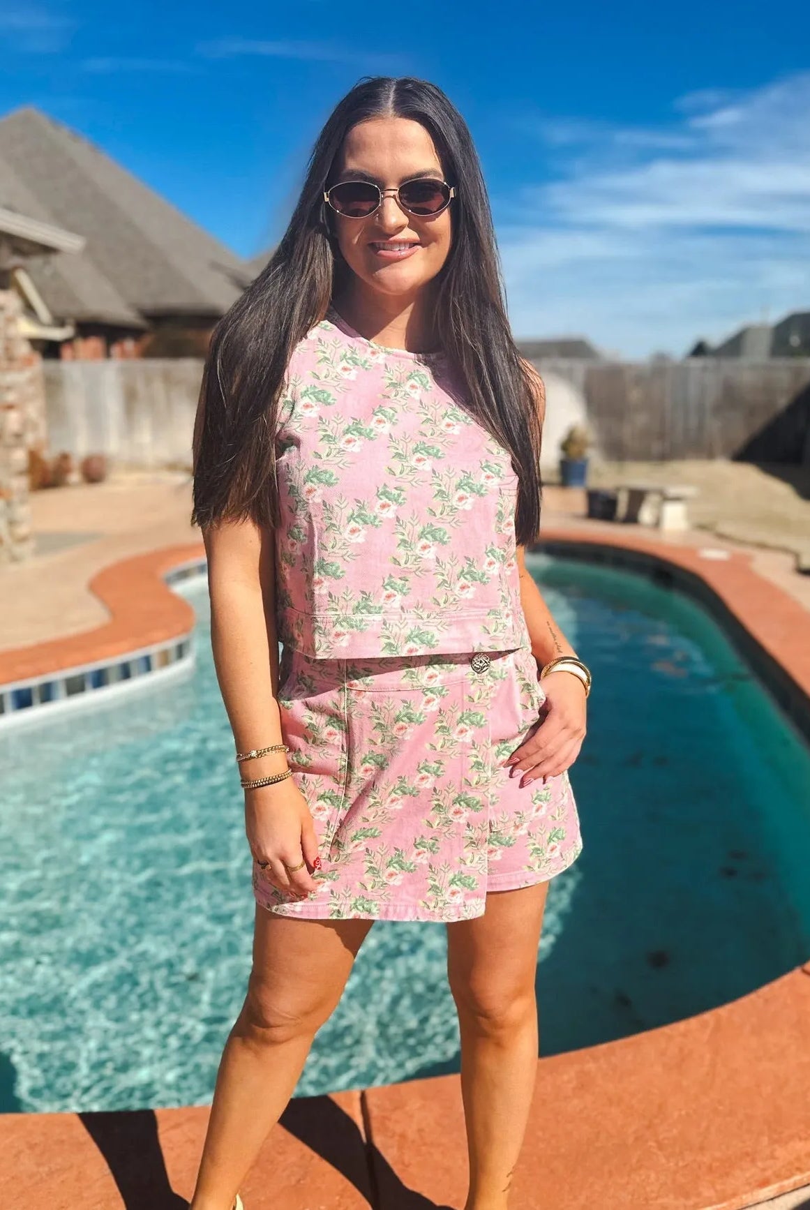Woman wearing a pink floral print denim skort and tank top set. She is smiling and wearing sunglasses outdoors by a pool.