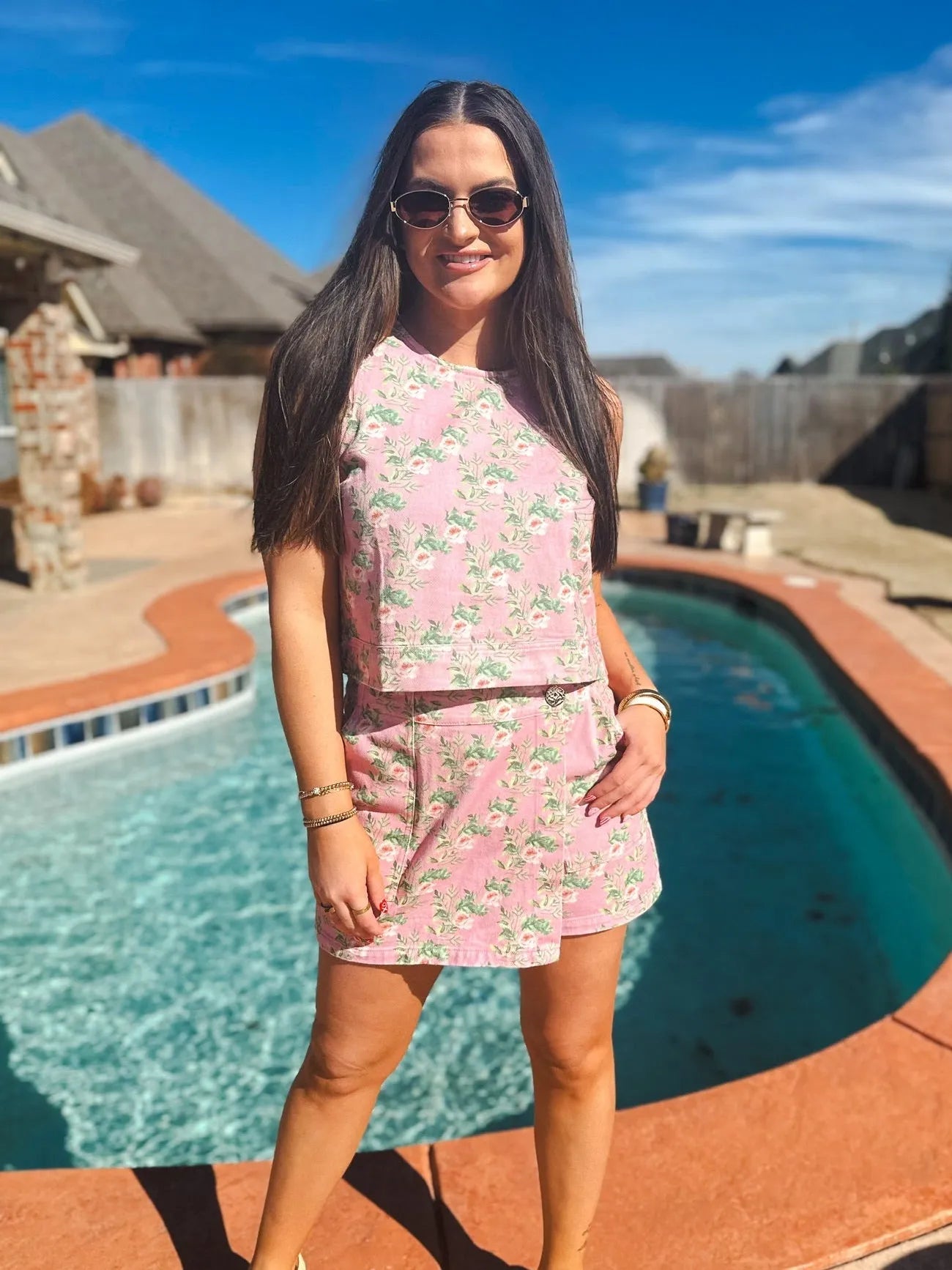 Woman wearing a pink floral print denim skort and tank top set. She is smiling and wearing sunglasses outdoors by a pool.