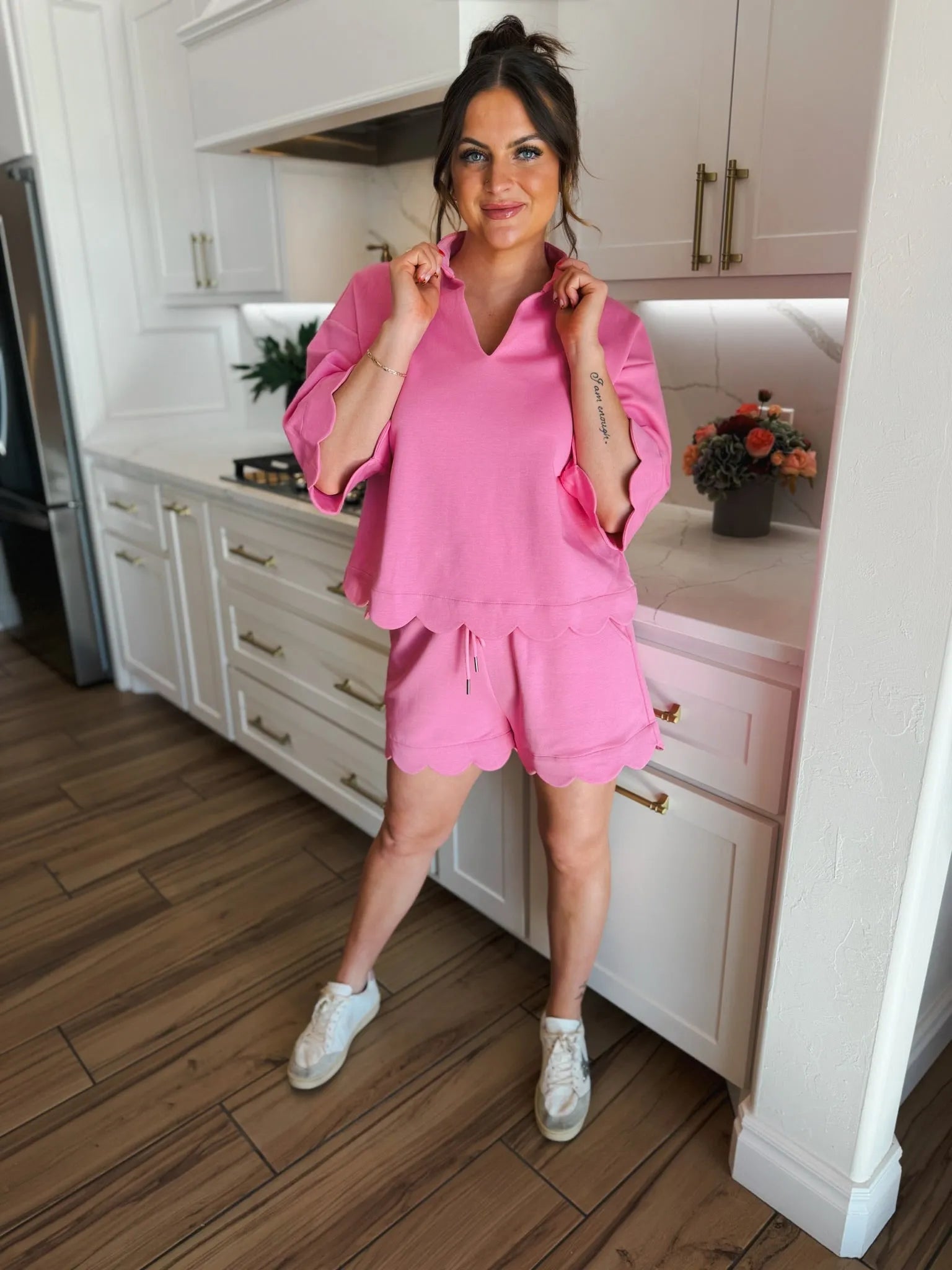 Woman wearing a pink scalloped short and top set by Entro, with matching white sneakers, standing in a kitchen.