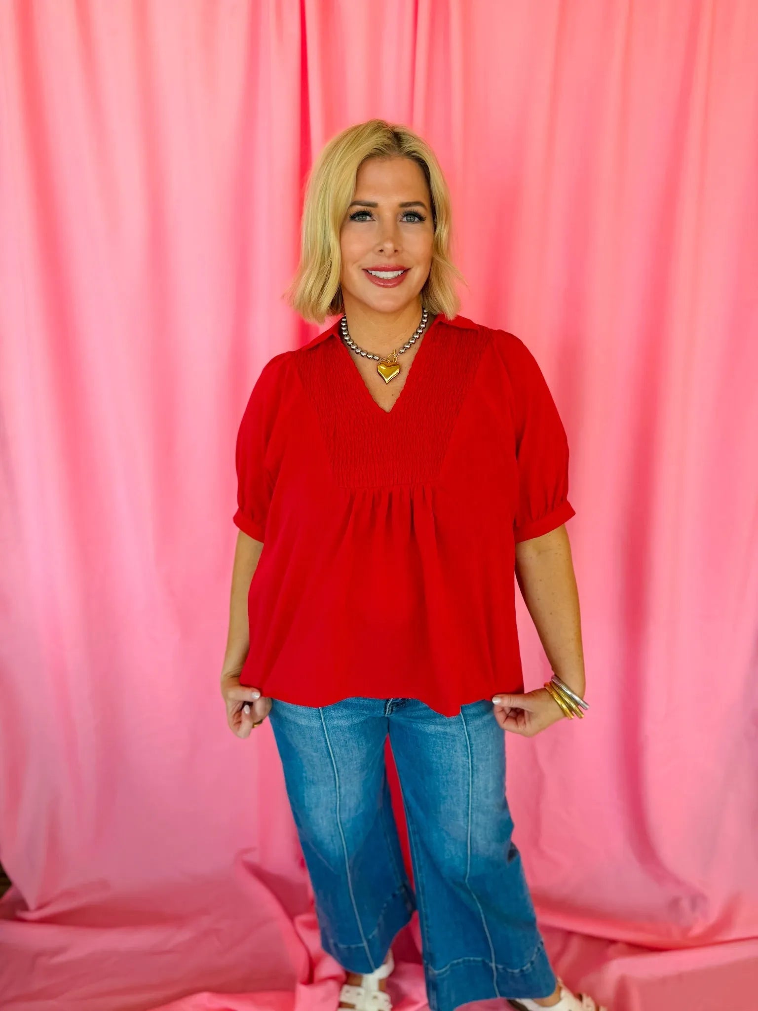 Entro red smocked V-neck blouse with short puff sleeves, paired with blue flared jeans. Woman wears heart necklace and