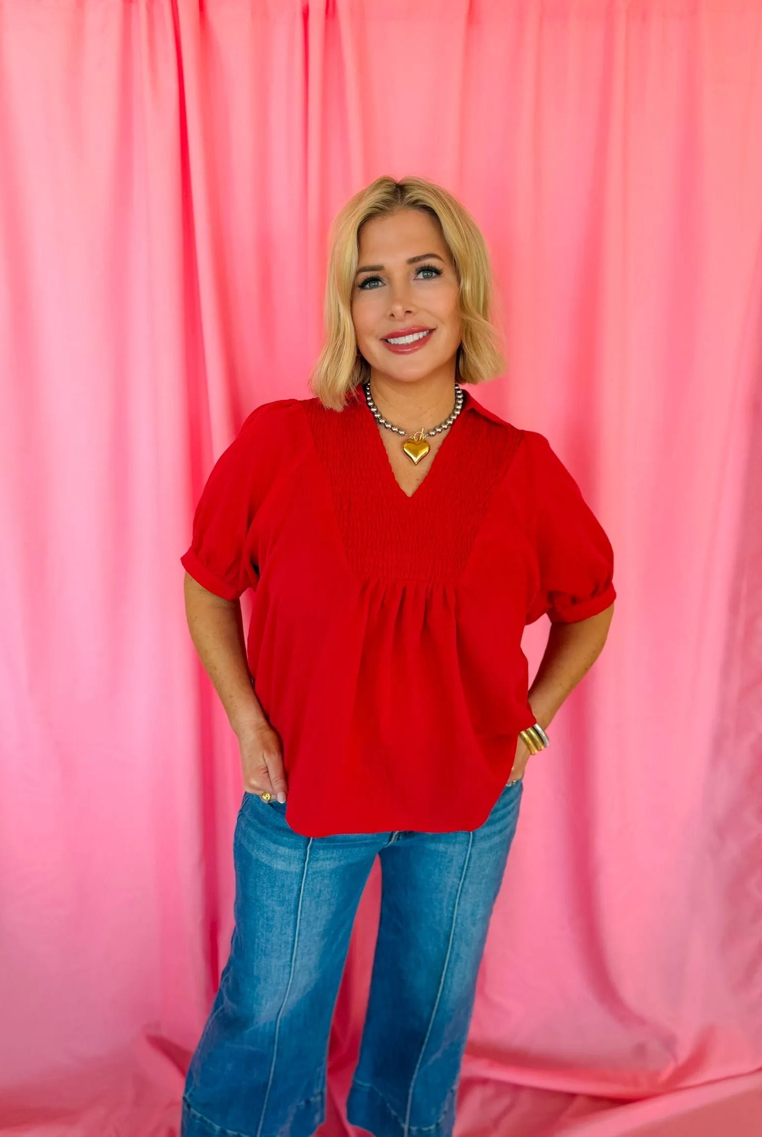 Woman wearing a red smocked V-neck blouse with puff sleeves and blue flared jeans, accessorized with a heart pendant