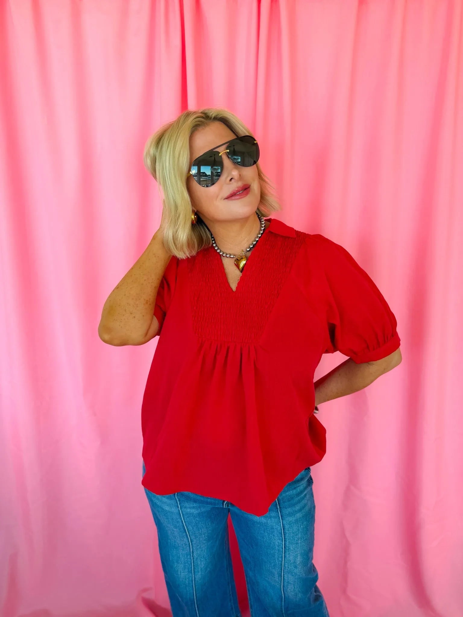 Woman wearing a red smocked V-neck blouse with short puff sleeves, blue jeans, and aviator sunglasses against a pink