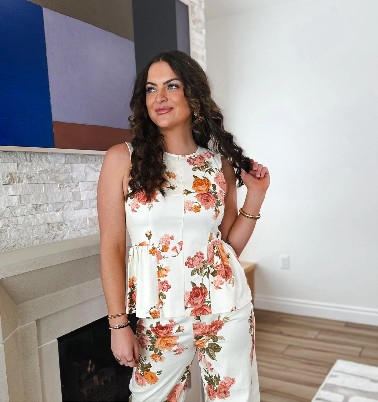 Woman in white floral print sleeveless peplum top and matching pants. Top has orange and pink roses pattern and zip back.