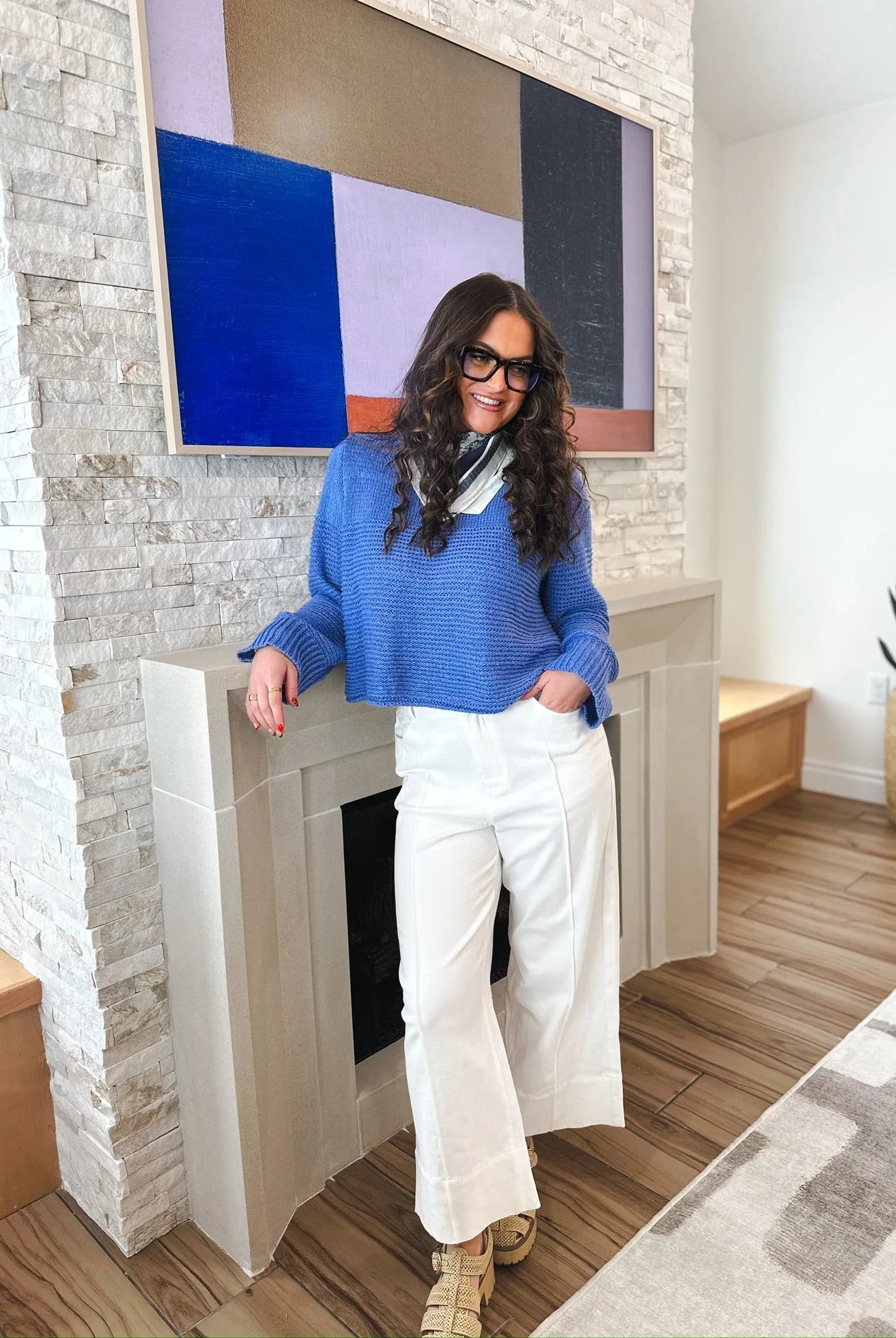 Woman in white wide-leg pleated jeans and a blue sweater, leaning against a fireplace. The jeans have a relaxed fit and a