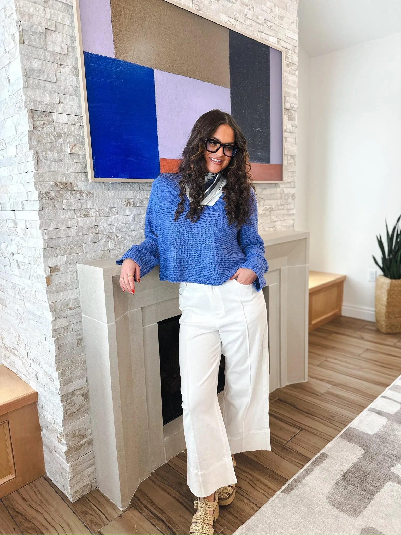 Woman in white wide-leg pleated jeans and a blue sweater, leaning against a fireplace. The jeans have a relaxed fit and a